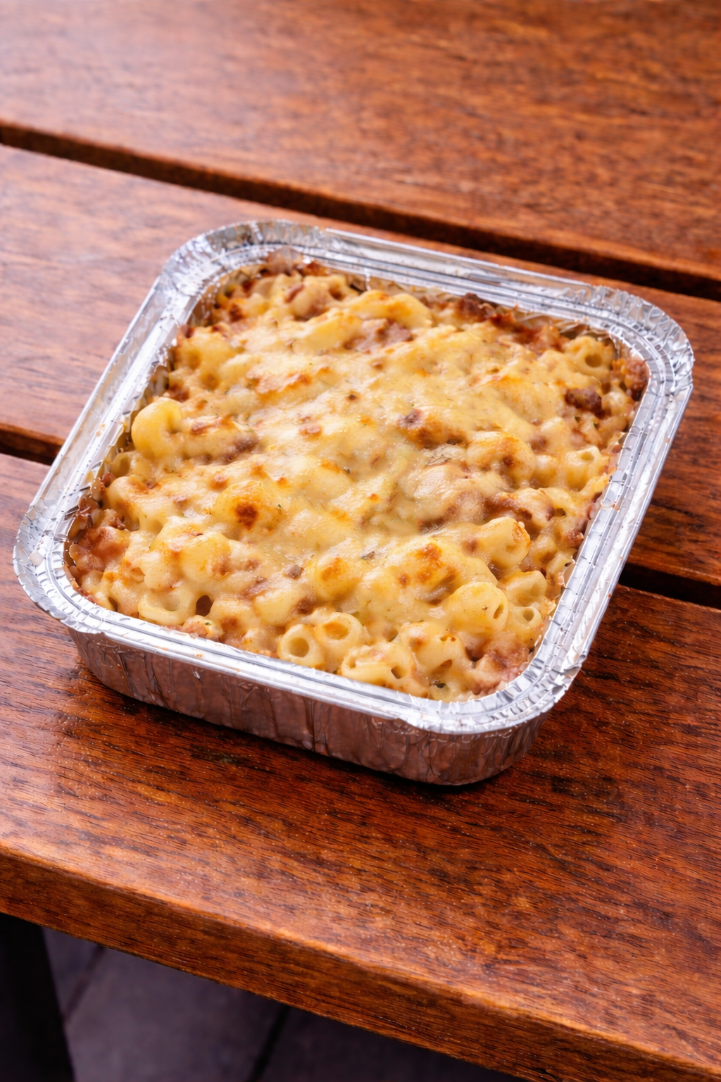 A foil container of baked macaroni and cheese with melted cheese on top, placed on a wooden table.