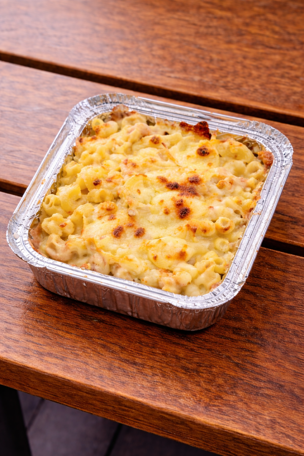 Baked macaroni and cheese in a foil container on a wooden table.