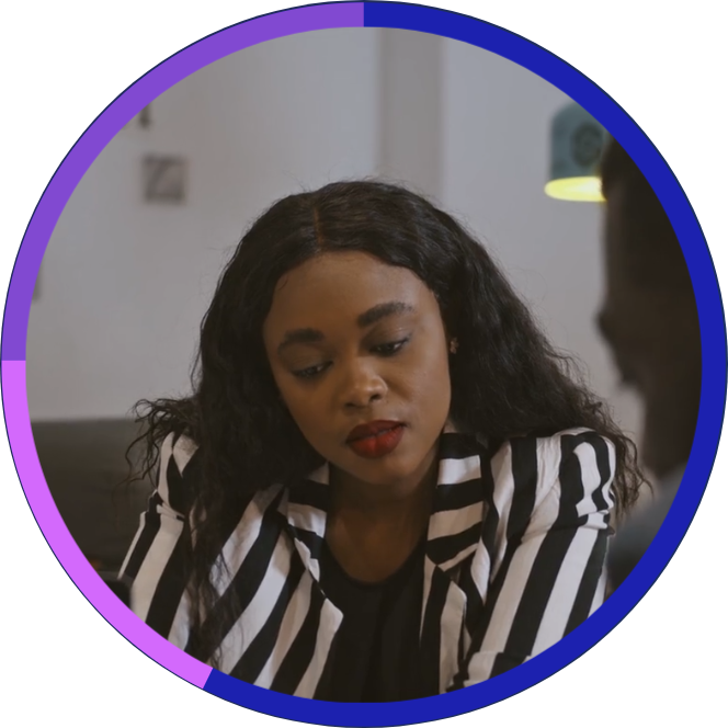 A woman with long, curly black hair and dark lipstick wearing a black and white striped blazer, talking to a person in a modern, well-lit room.
