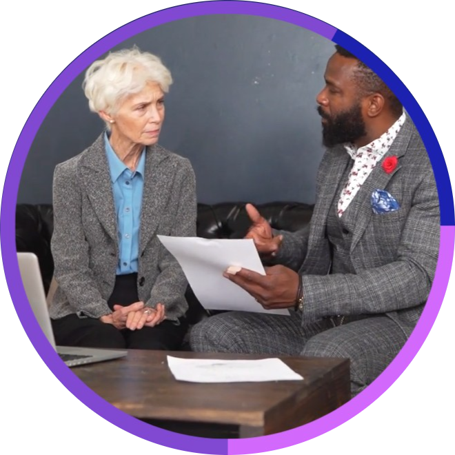 A woman with white hair and a man with a beard having a serious conversation during a meeting or interview, sitting on a black couch with papers on a wooden table in front of them. A show of Ally consults attention to solving your problems
