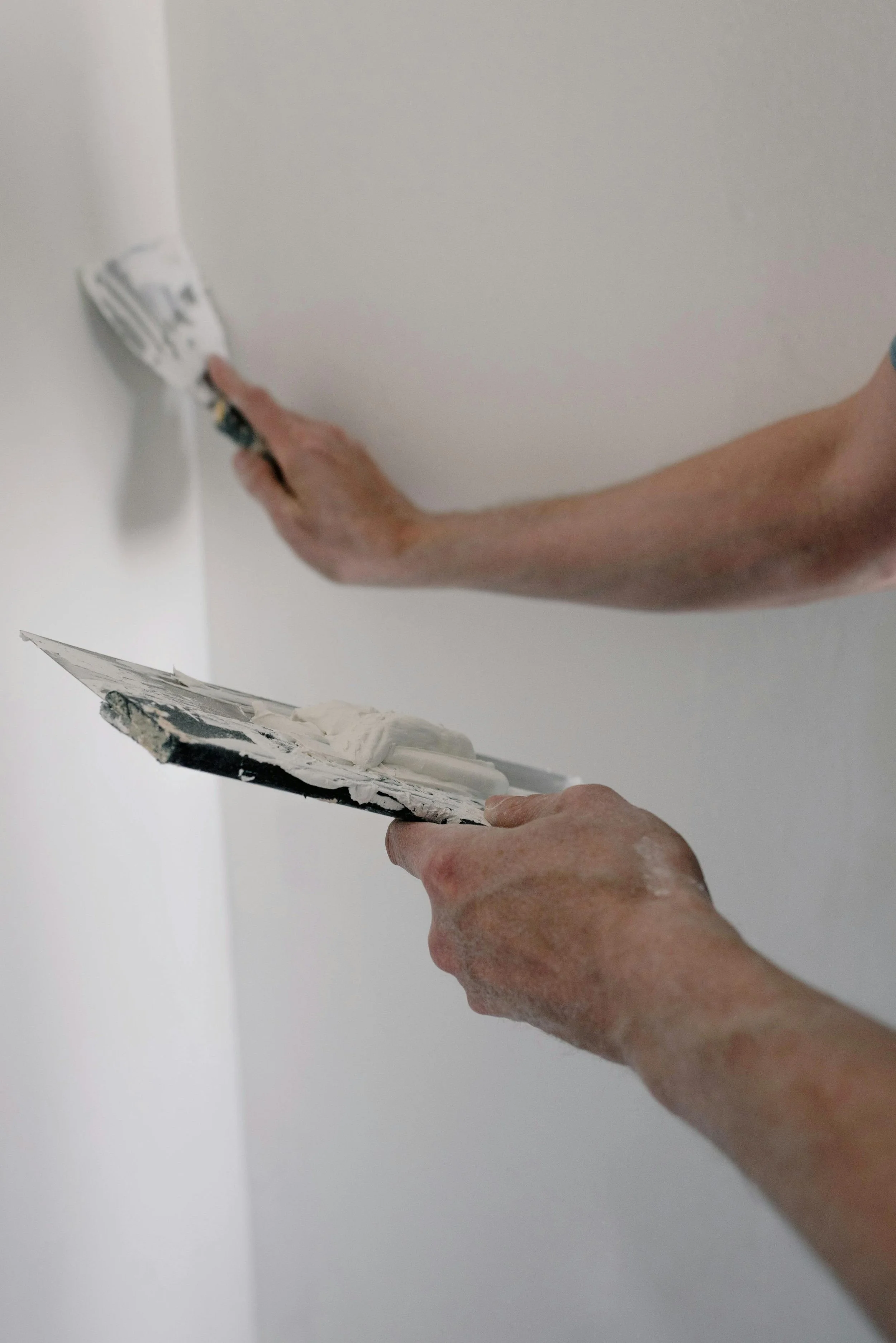 A person smoothing white paint on a ceiling with a putty knife, while holding a putty spatula in their other hand.