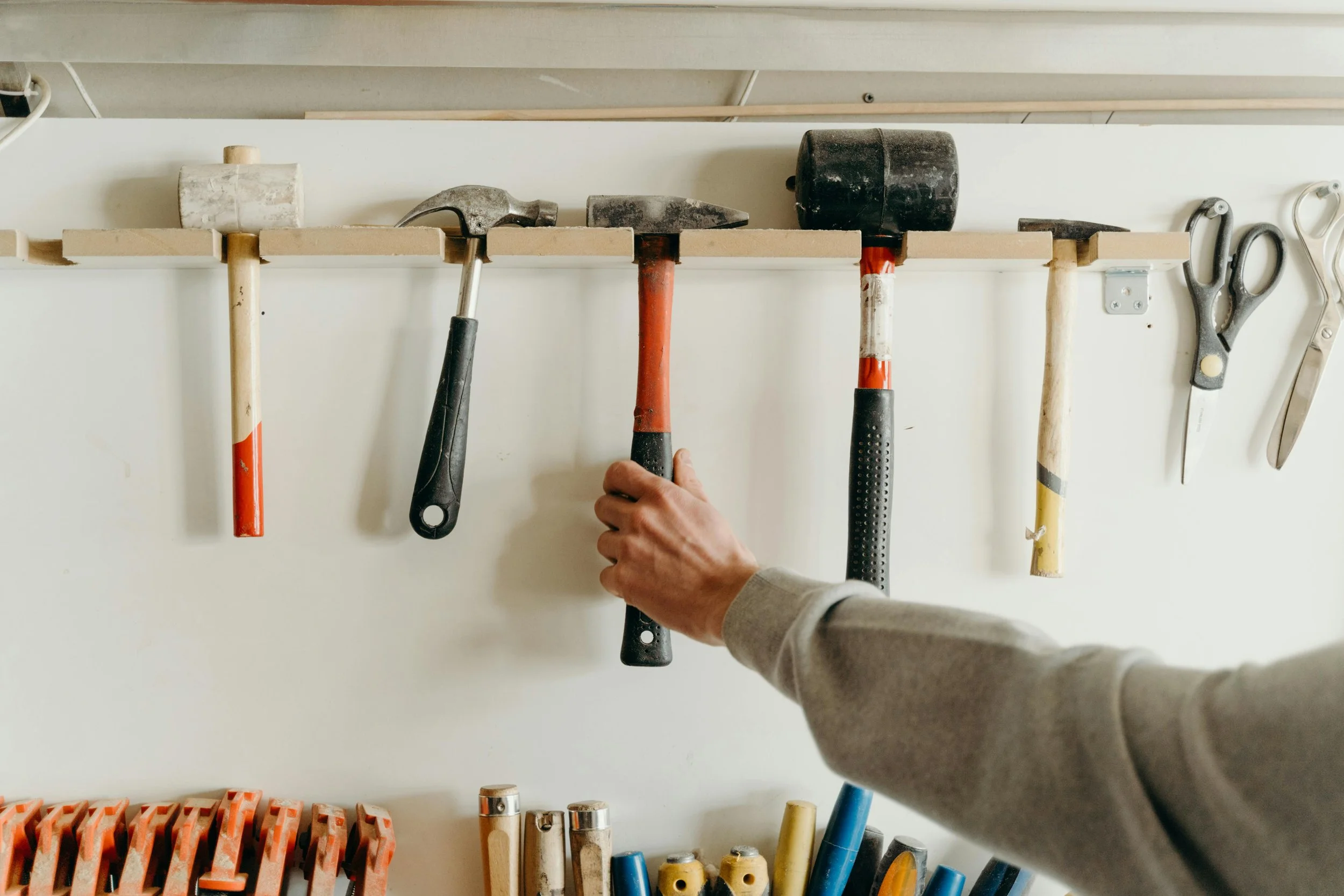 A person's hand hanging a hammer on a wooden wall rack with various tools, including screwdrivers and scissors, arranged on hooks.