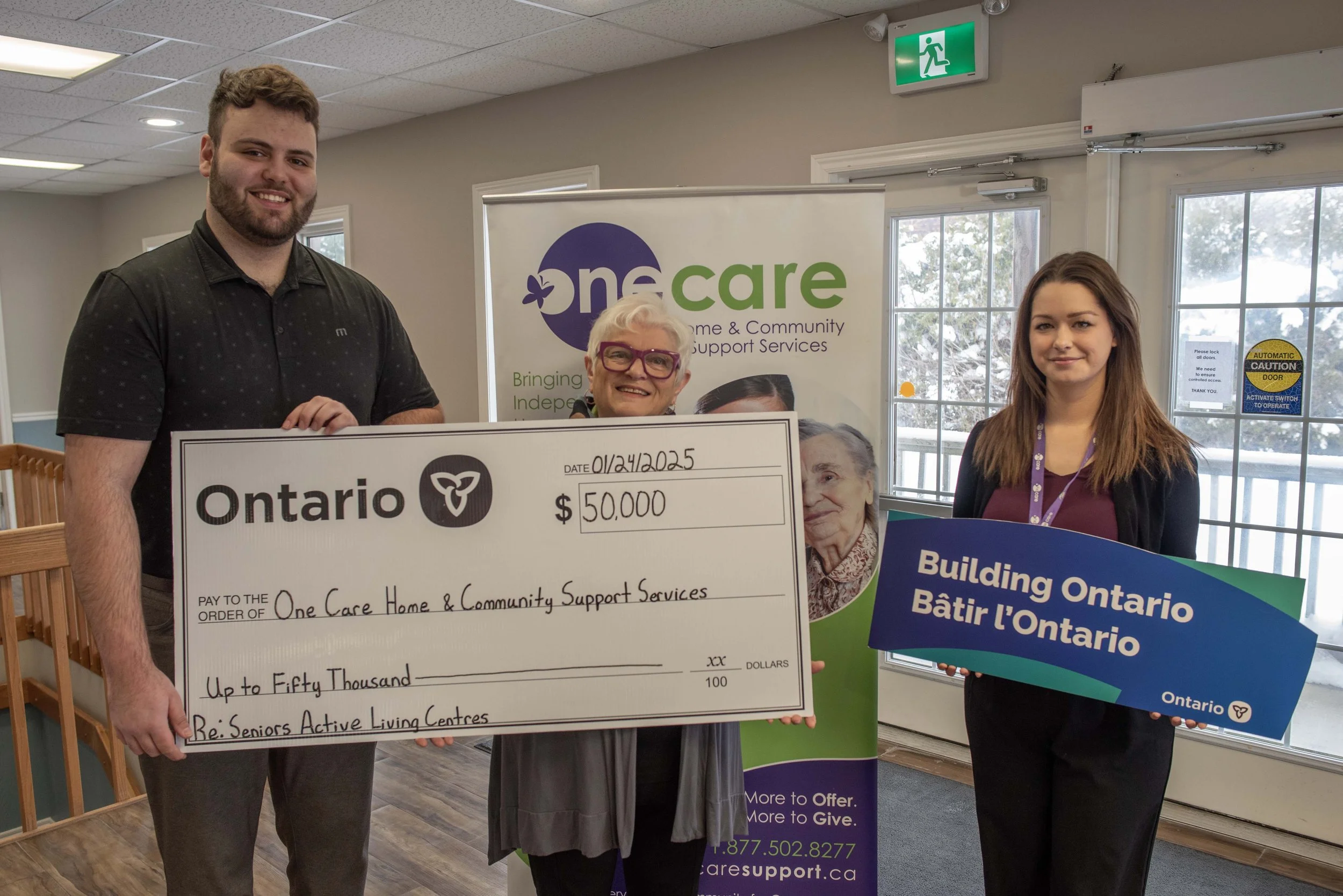 Three people standing indoors holding a large check and a sign at a donation event. The man on the left has curly hair, a beard, and is wearing a black shirt. The woman in the middle has white hair, glasses, and is smiling. The young woman on the right has long brown hair and is holding a blue and green sign. Behind them, a banner displays the words 'One Care' and a logo, indicating a support service organization. There is an exit sign above the door and large windows in the background.
