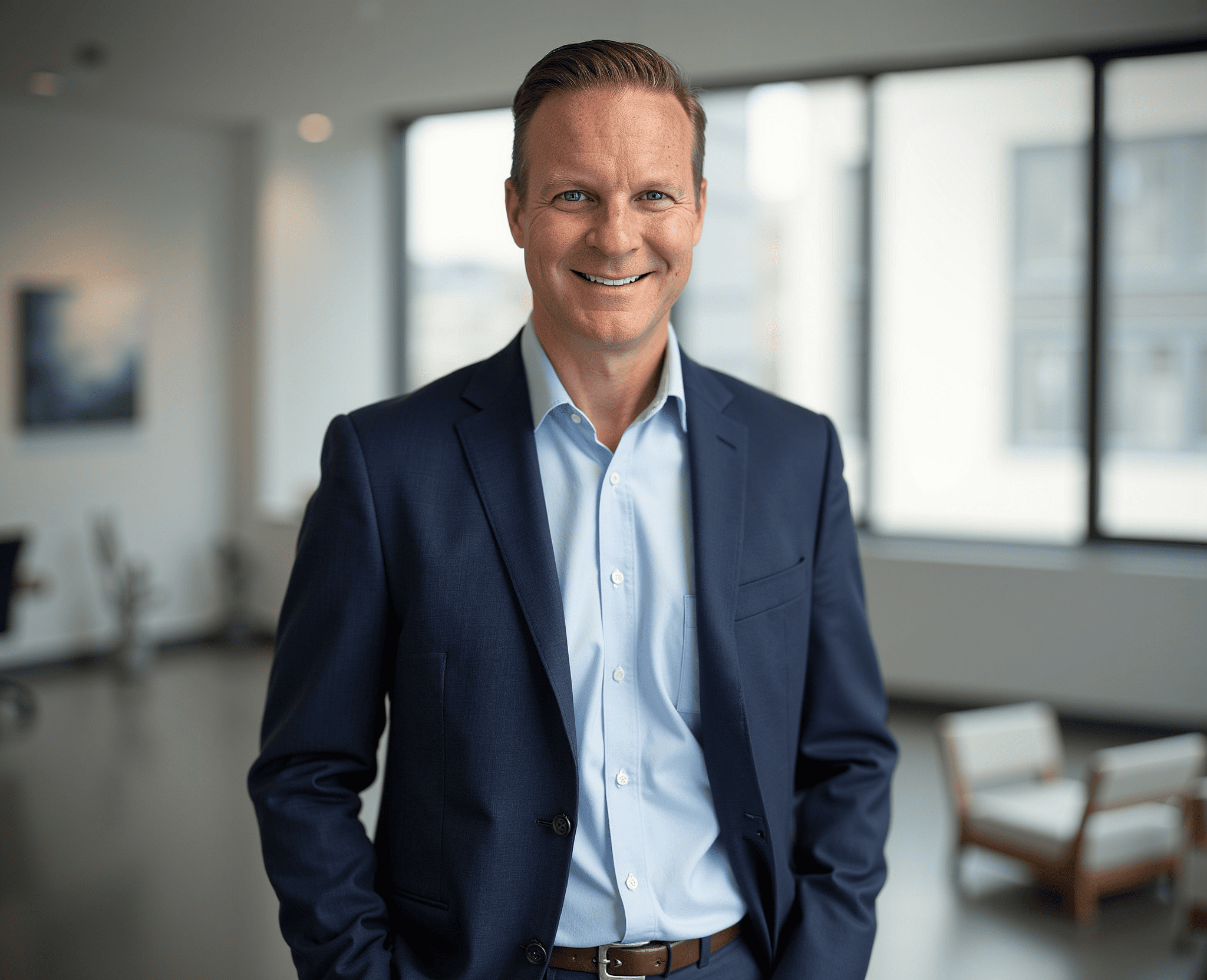 A smiling man in a dark blue suit and light blue shirt standing in an office with large windows and modern furniture.