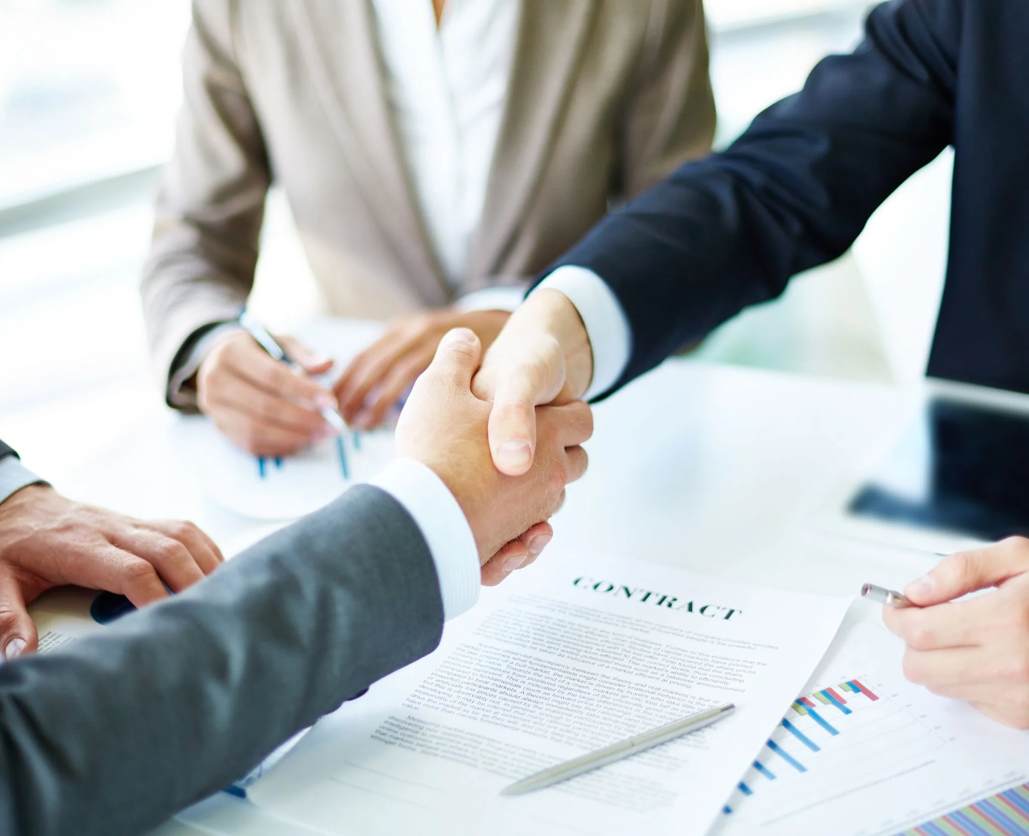Two businesspeople shaking hands over a table with a contract document, charts, and a pen.