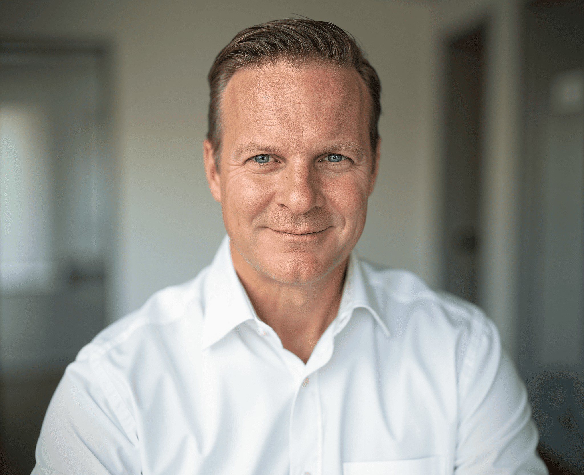 A middle-aged man with short light brown hair, wearing a white button-down shirt, smiling and looking directly at the camera in an indoor setting.