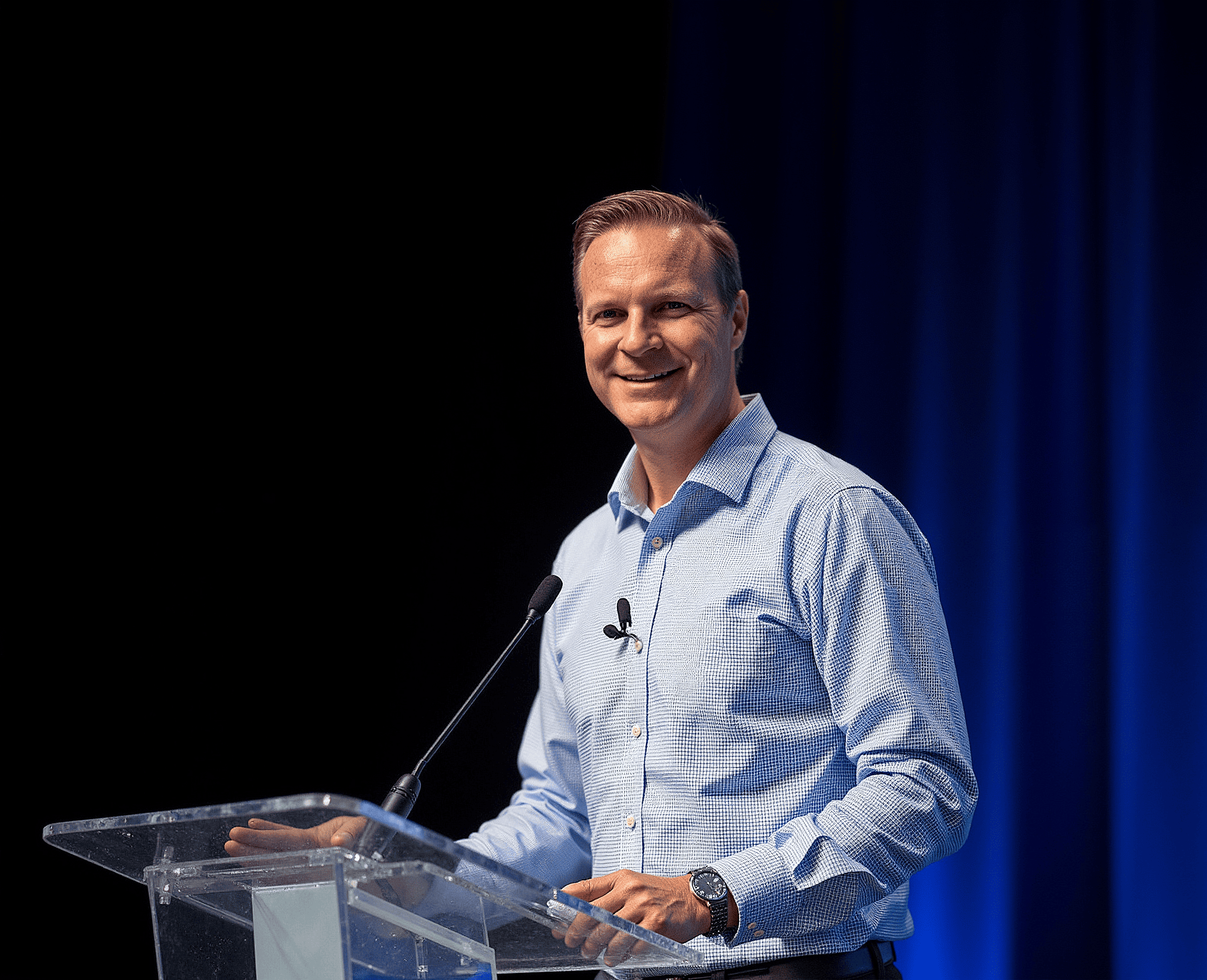 Man in a light blue dress shirt speaking at a podium with a microphone, smiling,perspective portrait, dark background with blue curtains.