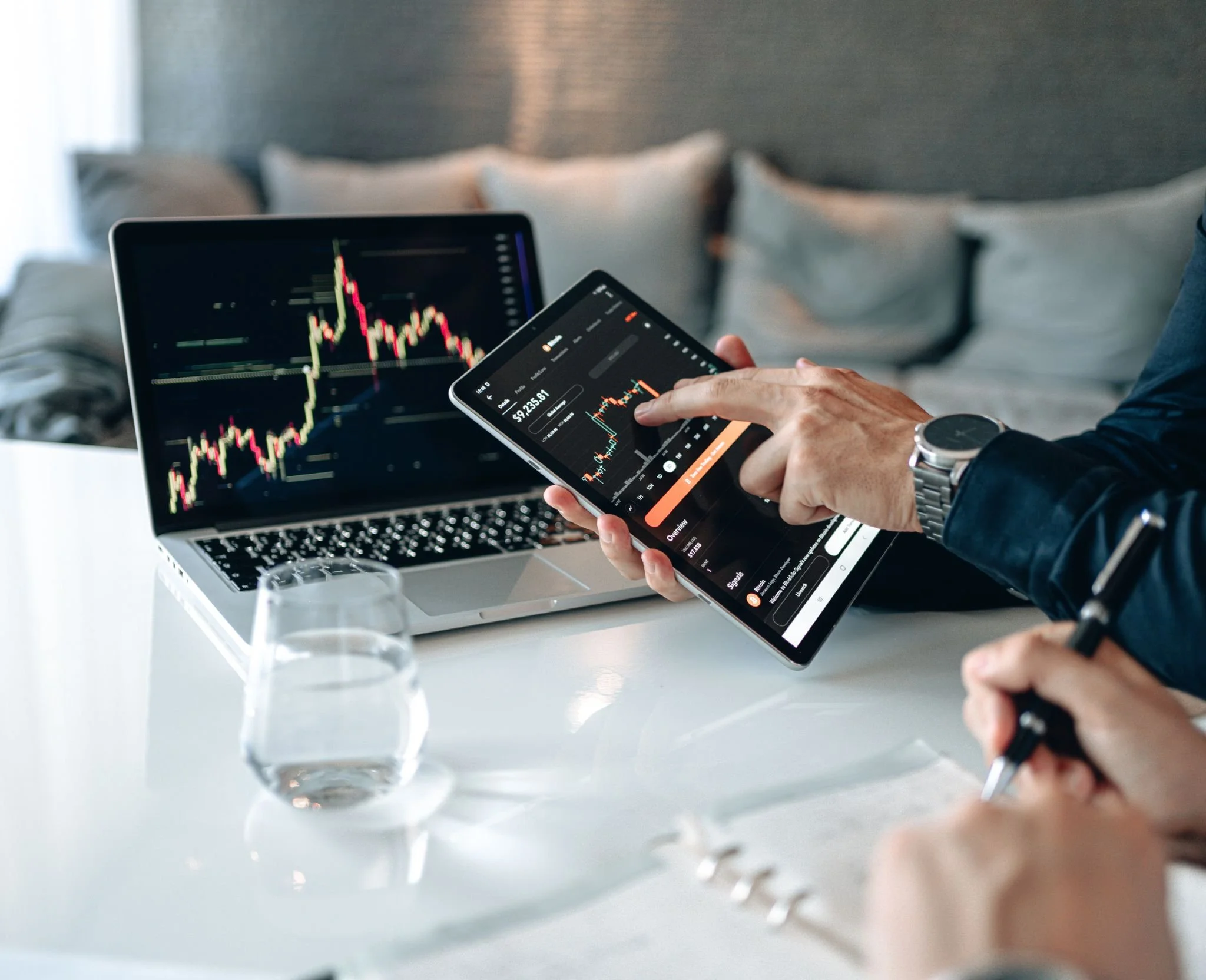 Person using a tablet to analyze stock charts with a laptop displaying financial data on a table.
