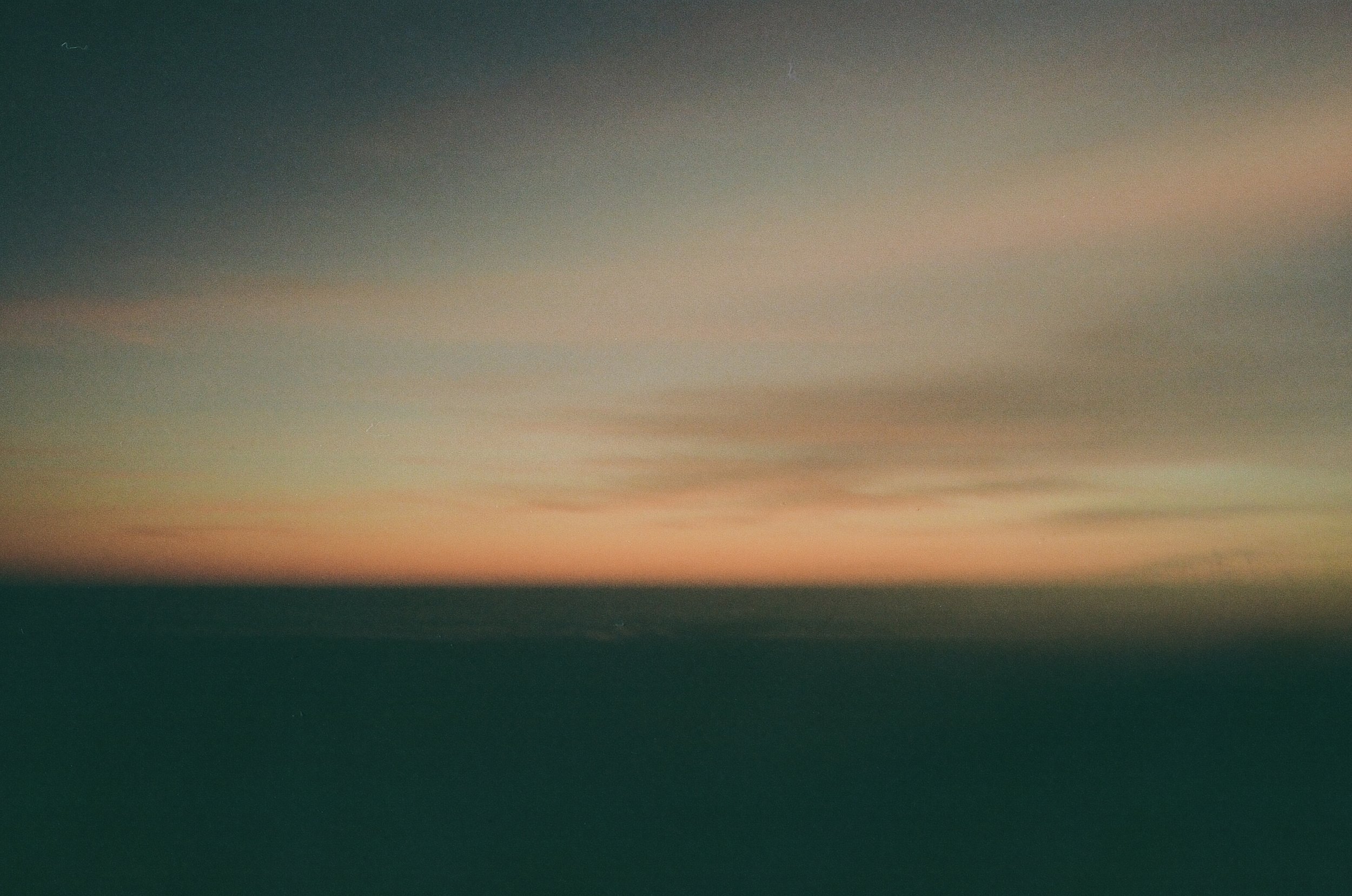 A blurred photograph of a sunset or sunrise over the ocean, with soft pastel colors and a dark foreground.