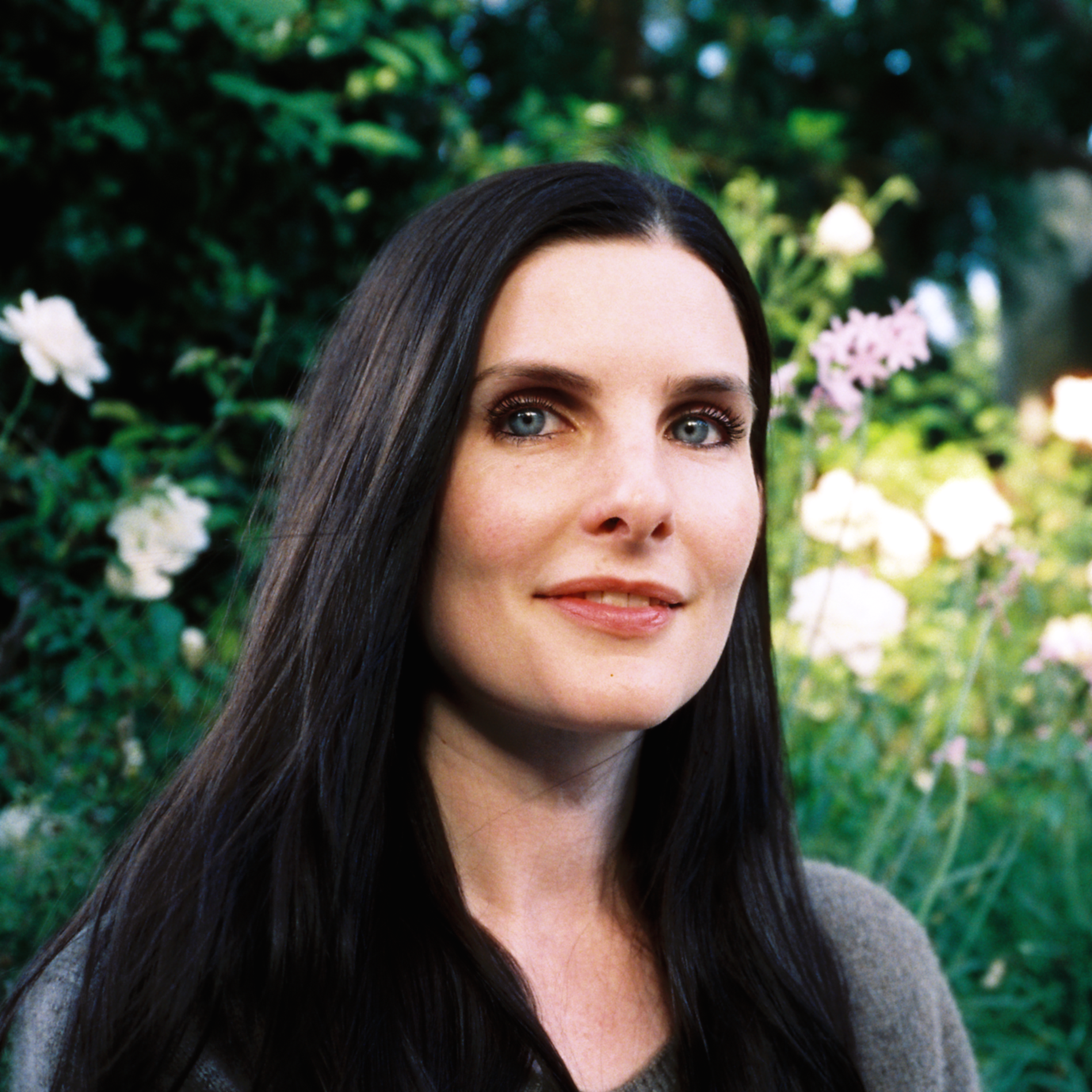A woman with long black hair and blue eyes smiling outdoors surrounded by blooming white and pink flowers and green foliage.