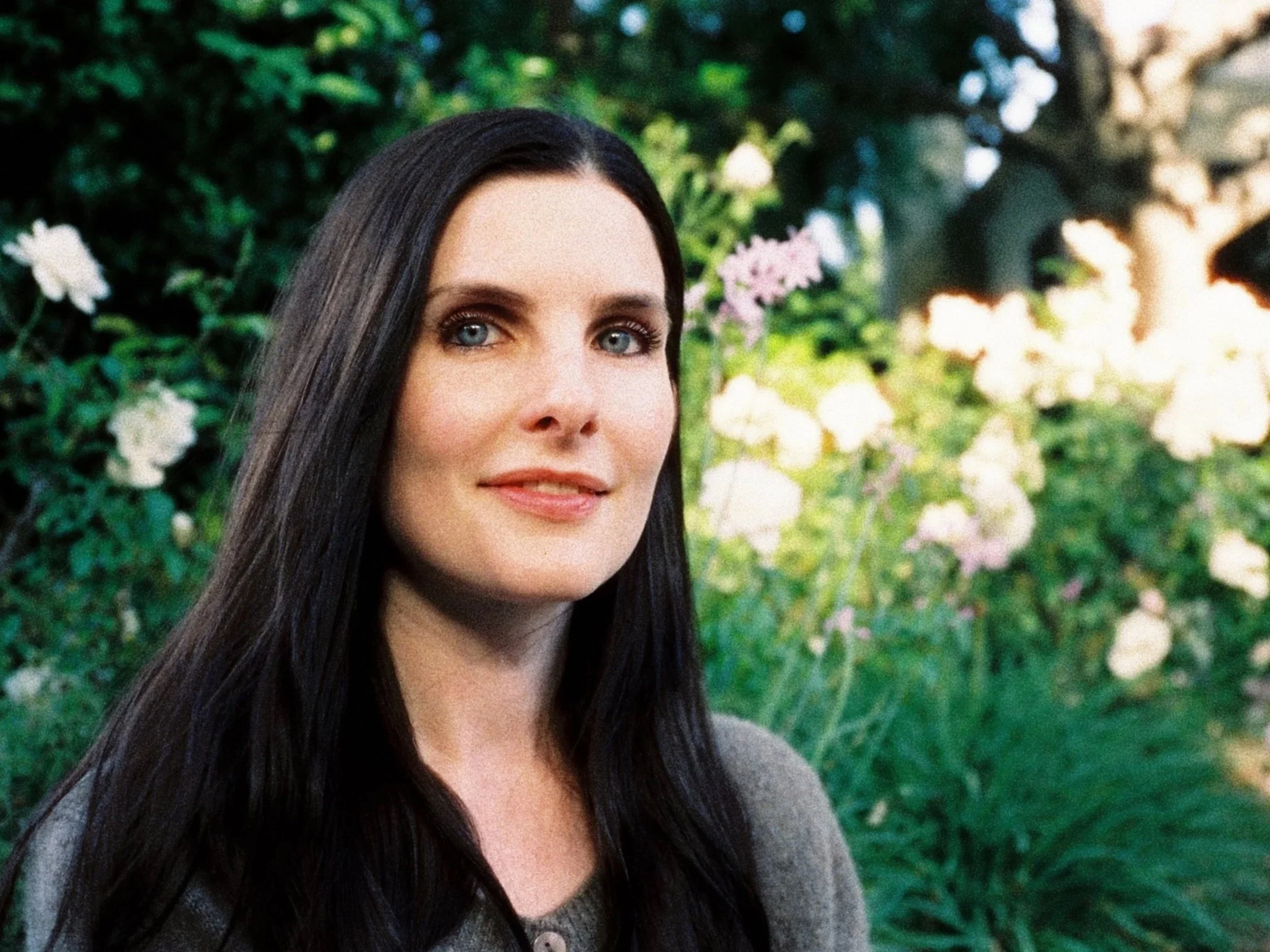 A woman with long dark hair and blue eyes stands outdoors in front of green foliage and flowers.
