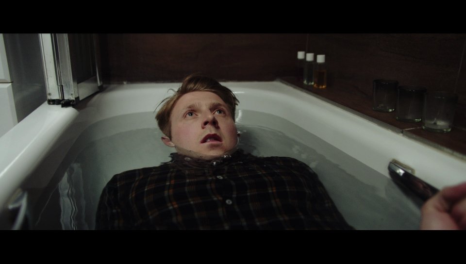 A person in a plaid shirt lying in a bathtub filled with water, looking upwards. The bathtub is against a wall with bottles on the ledge.