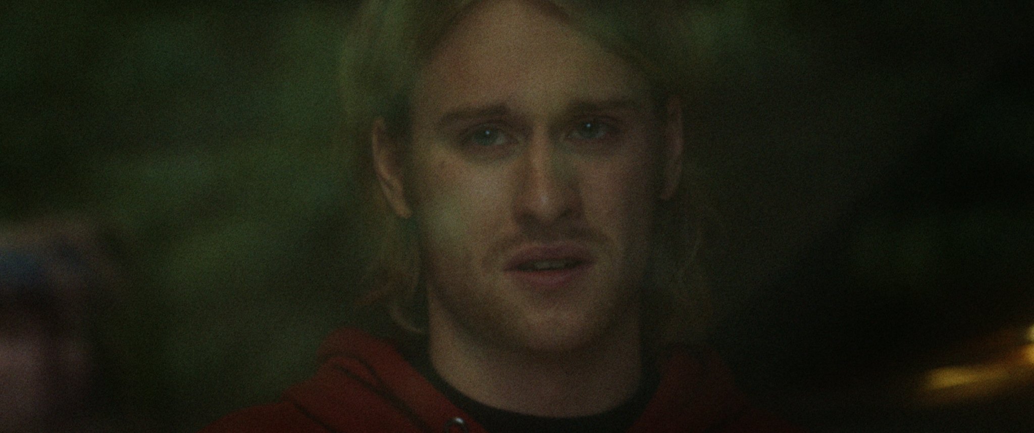 Close-up of a person with long hair in a red hoodie, looking through glass, with a blurred dark background.