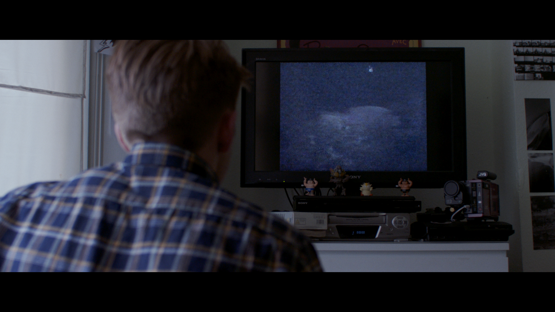 Person in a plaid shirt watching a fuzzy black-and-white display on a TV, with figurines and electronic devices nearby.