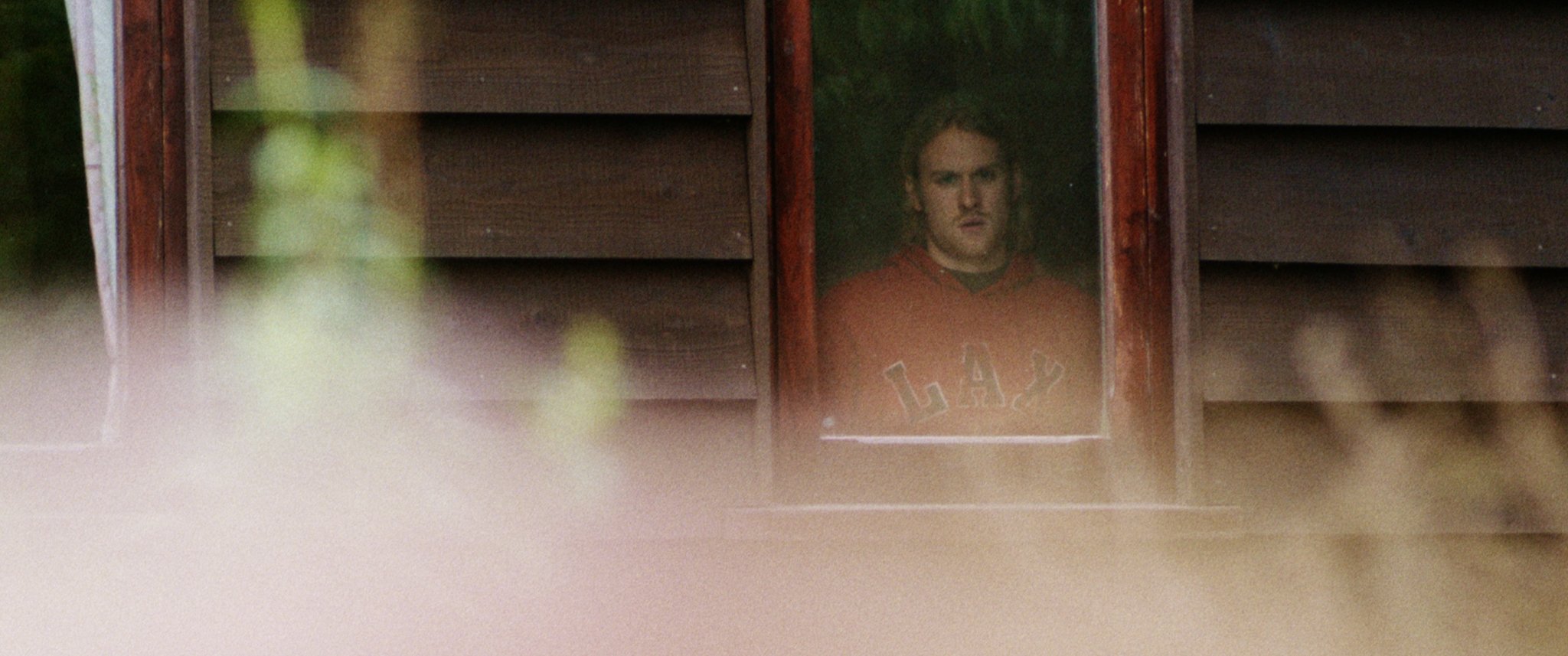 A person wearing a red hoodie is looking out from a window of a wooden building. The foreground is blurred with plants.