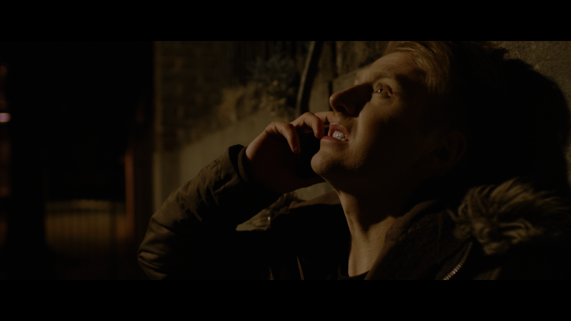 A person with blonde hair is leaning against a wall at night, holding a phone to their ear and looking upward, with a serious expression. They are wearing a dark fur-collared jacket.