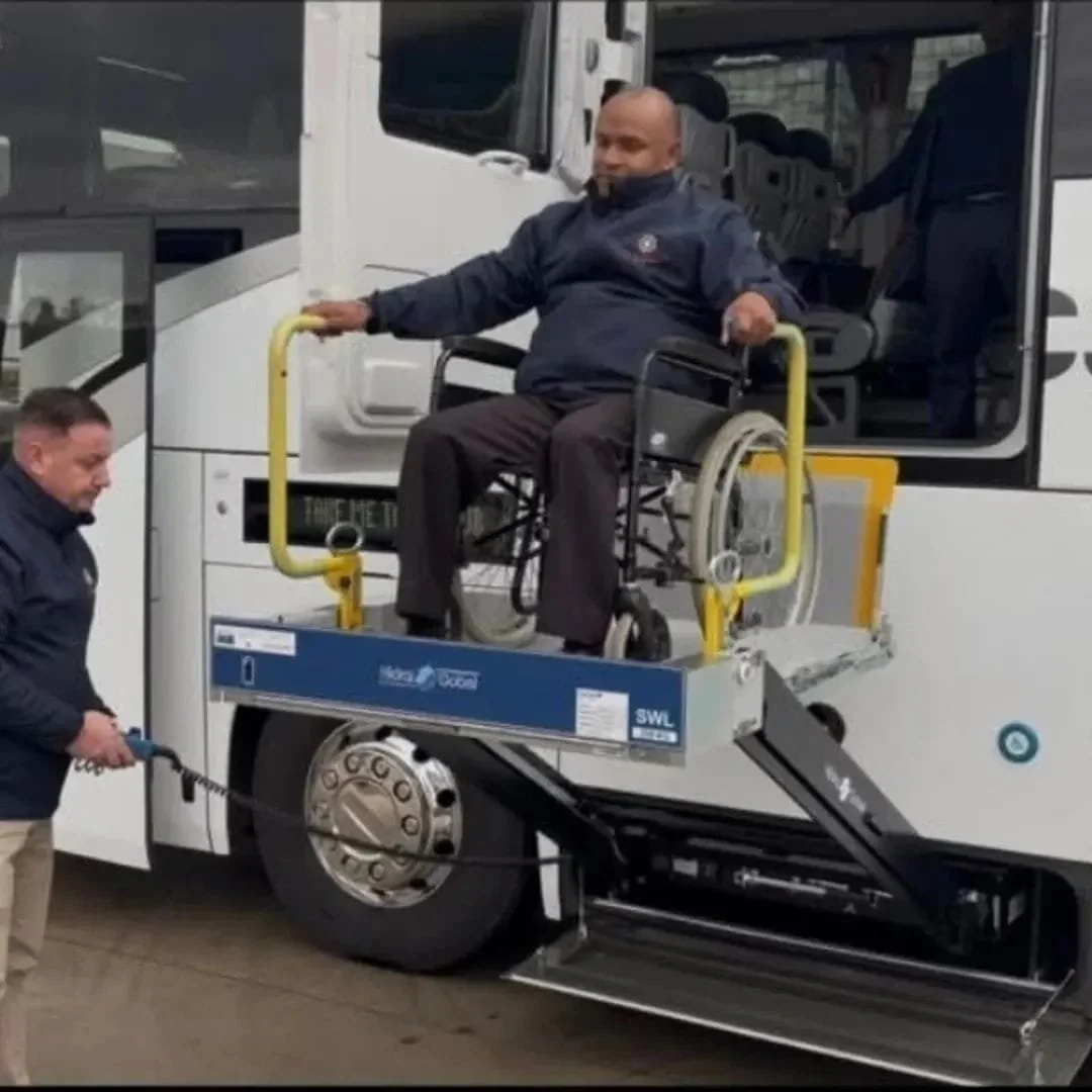A person in a wheelchair being lowered onto a bus using a wheelchair lift operated by another person.