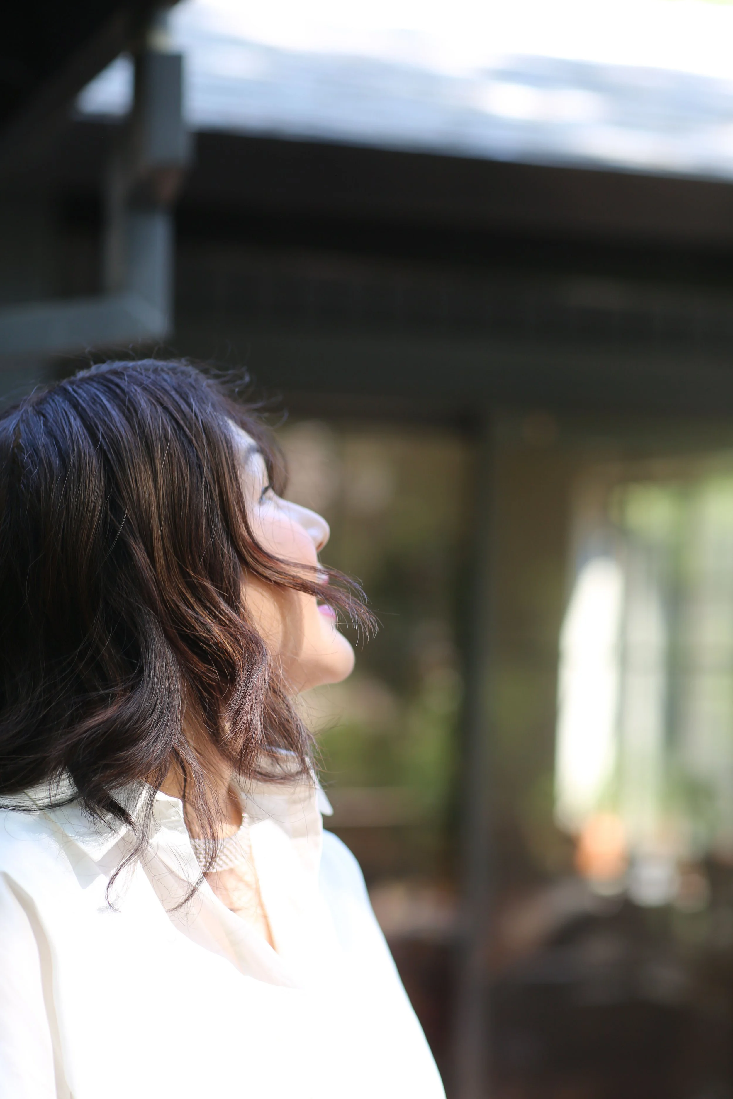 Side profile of a woman with wavy dark hair looking to the left, wearing a white shirt, in natural daylight with blurred background.