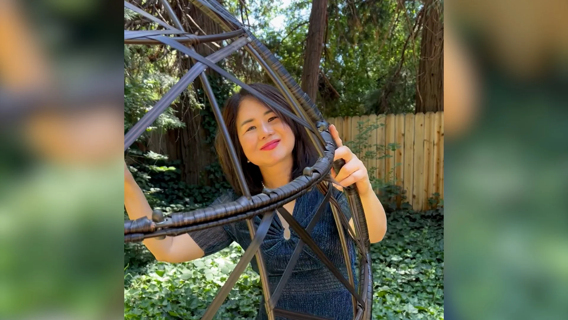 Woman smiling and playing with a large wicker fire pit or decorative structure outdoors in a garden with trees and a wooden fence in the background.