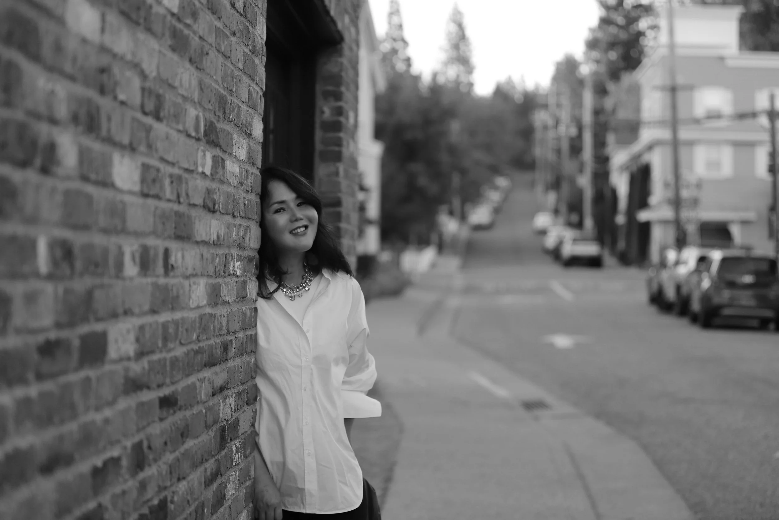 A woman with dark hair and a necklace, leaning against a brick wall on a city street, smiling at the camera in black and white.