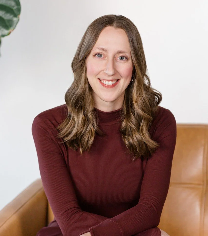 A woman with wavy brown hair, wearing a burgundy long-sleeve top, smiling and sitting on a tan leather chair, with a white background and a partial green plant visible on the left.