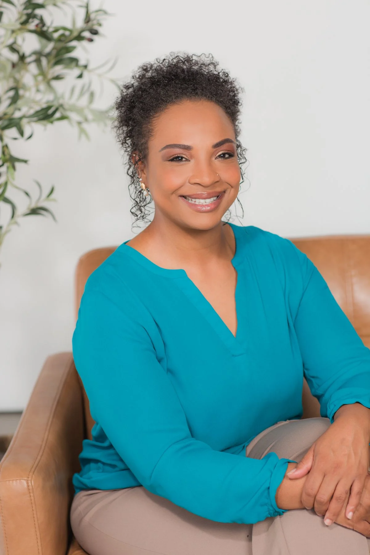 A psychotherapist smiling, sitting on a tan chair with her arms crossed, wearing a teal blouse, with a background of a white wall and a green plant.