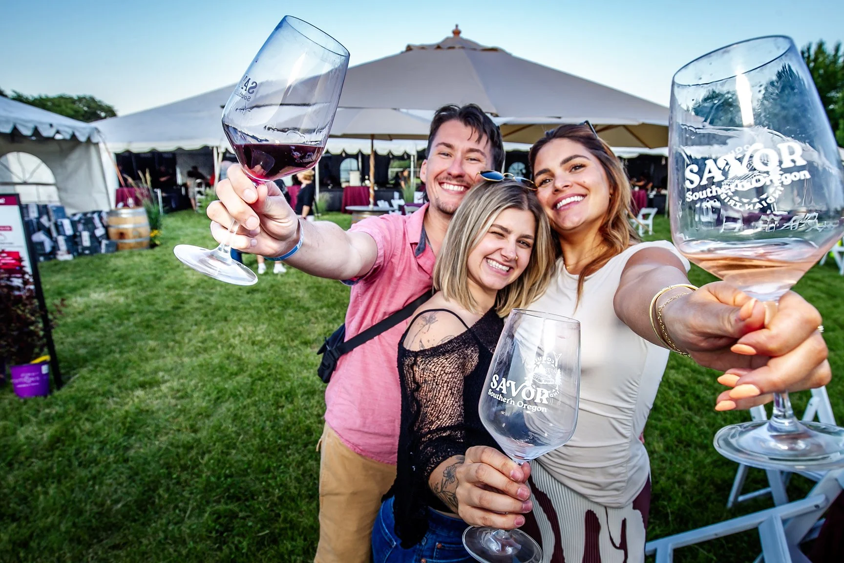 Three friends smiling and taking a selfie with wine glasses at an outdoor event in Southern Oregon.