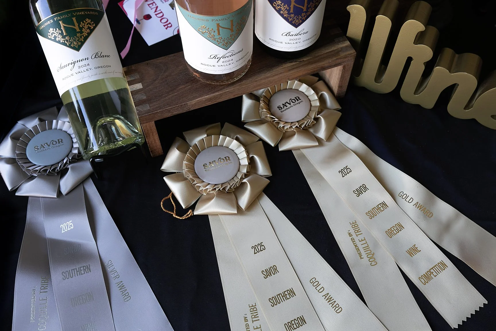 Wine bottles with labels from Haume's Family Vineyards and related wines, several award ribbons with ribbons dated 2025, and a decorative wooden display on a black surface.