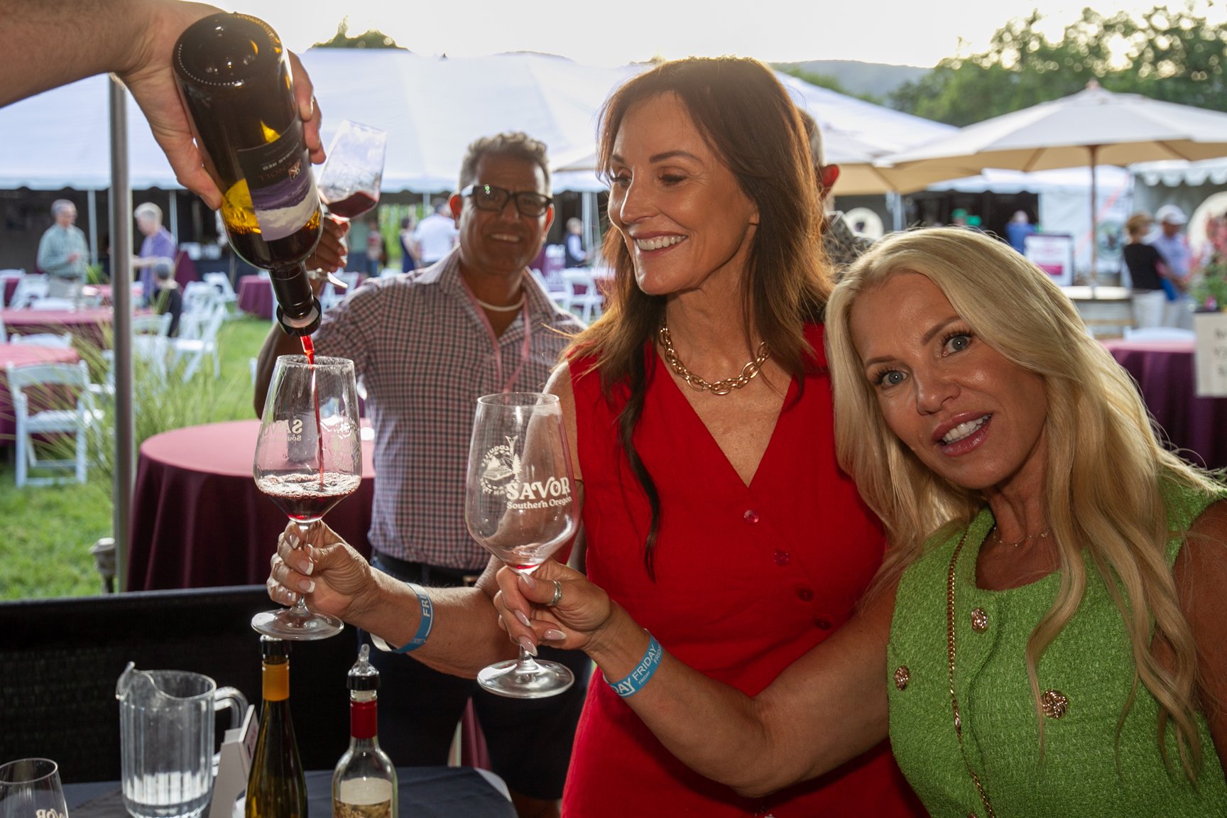 Two women at an outdoor event holding wine glasses, with a server pouring red wine into one of the glasses. A man stands in the background, smiling, with other tables and people visible under tents.