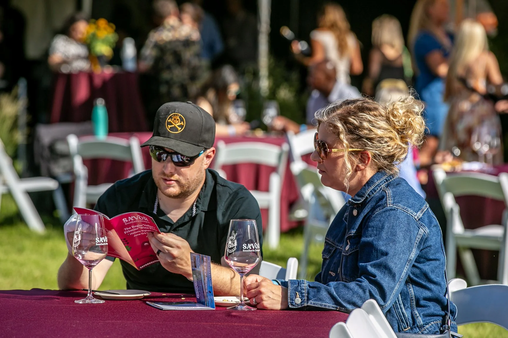 A man and woman sitting at a table outdoors, looking at a menu, with wine glasses in front of them. The man wears sunglasses, a black cap with a logo, and a black shirt. The woman has curly blonde hair, sunglasses, and a denim jacket. In the background, there are other people, some standing, around tables with burgundy tablecloths.