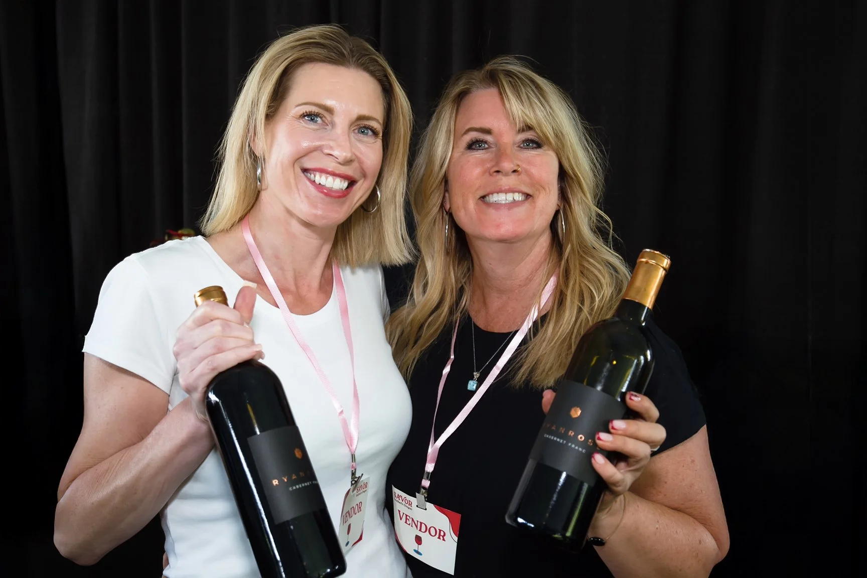 Two smiling women holding bottles of wine, wearing vendor badges, standing in front of a black background.