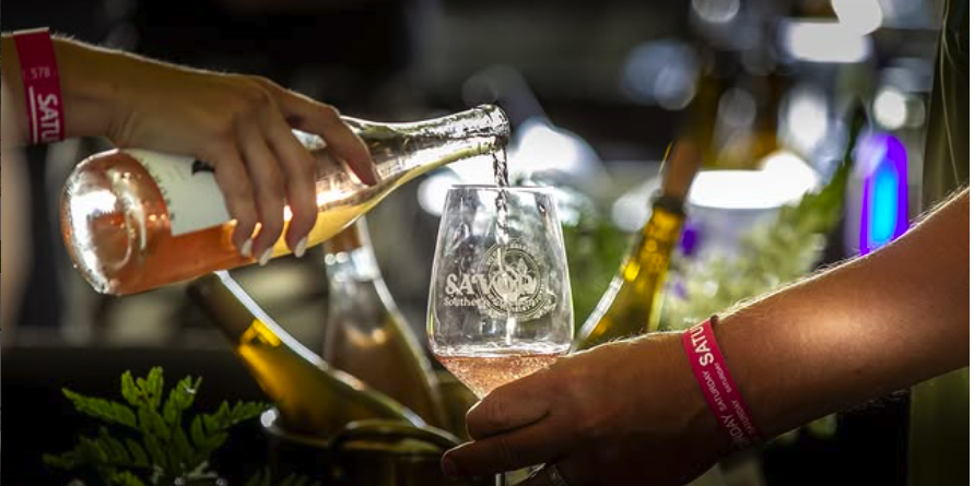 Person pouring rosé wine into a glass at a bar or wine shop, with bottles and green foliage in the background.