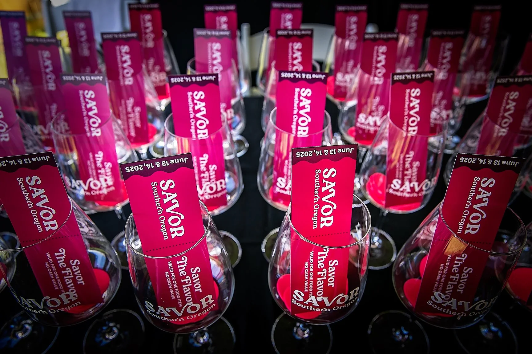 Multiple wine glasses with pink and black labels reading "SAVOR" and "Southern Oregon," arranged on a black surface, each containing red heart-shaped candies.