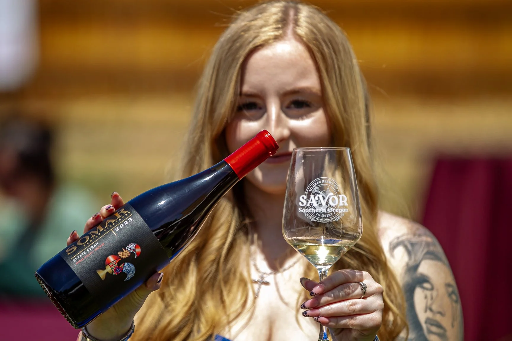 A woman with long red hair pouring white wine from a bottle into a wine glass. The wine bottle has a black and red label. The woman is smiling and holding the glass, which has a logo that reads 'SAVOR Southern Oregon.' Her nails are painted and she has tattoos on her arm.