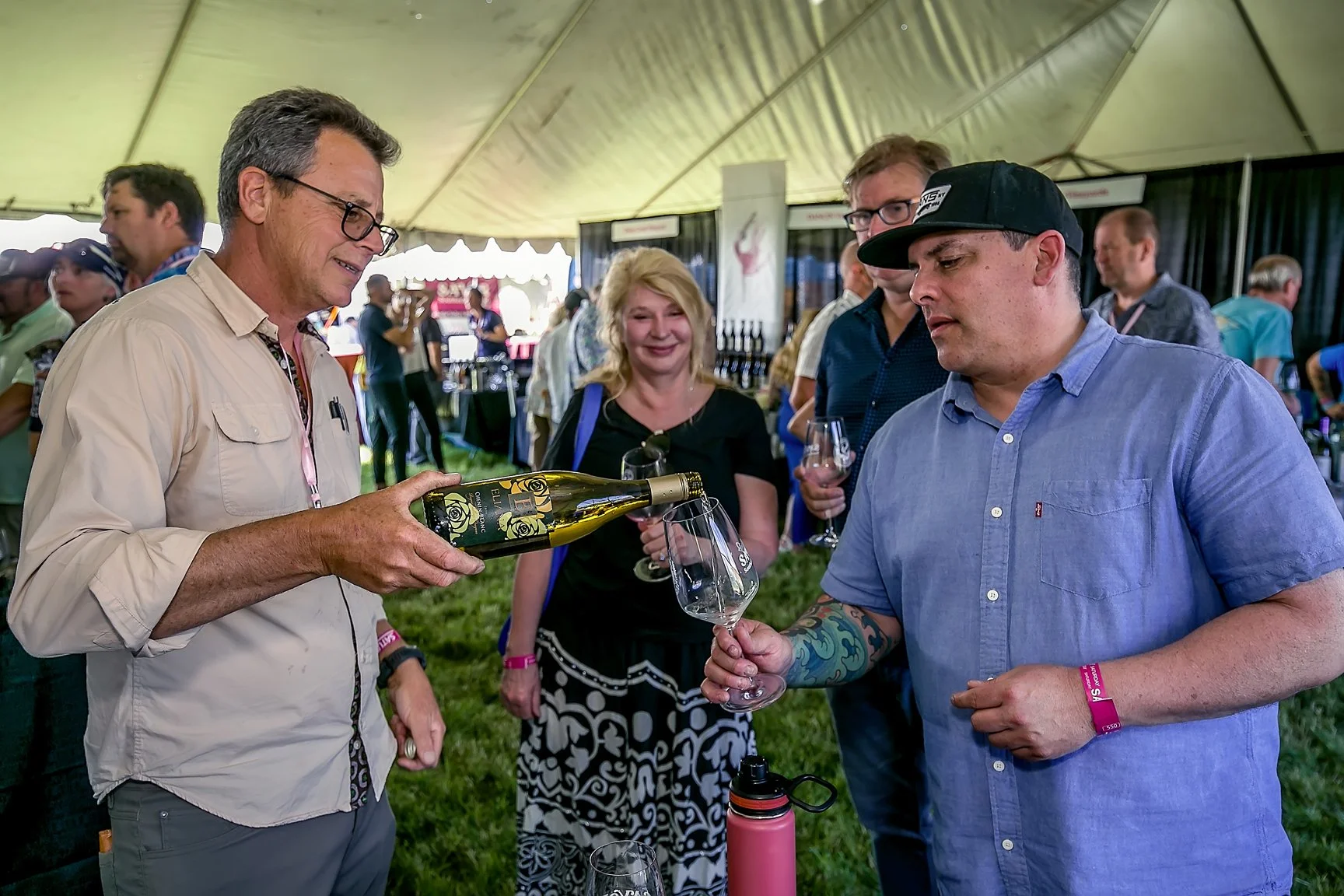 A man in a beige shirt and glasses pours wine into a glass held by a man in a blue shirt with tattoos and a black cap, while women and other people stand in a tented area at a wine tasting event.