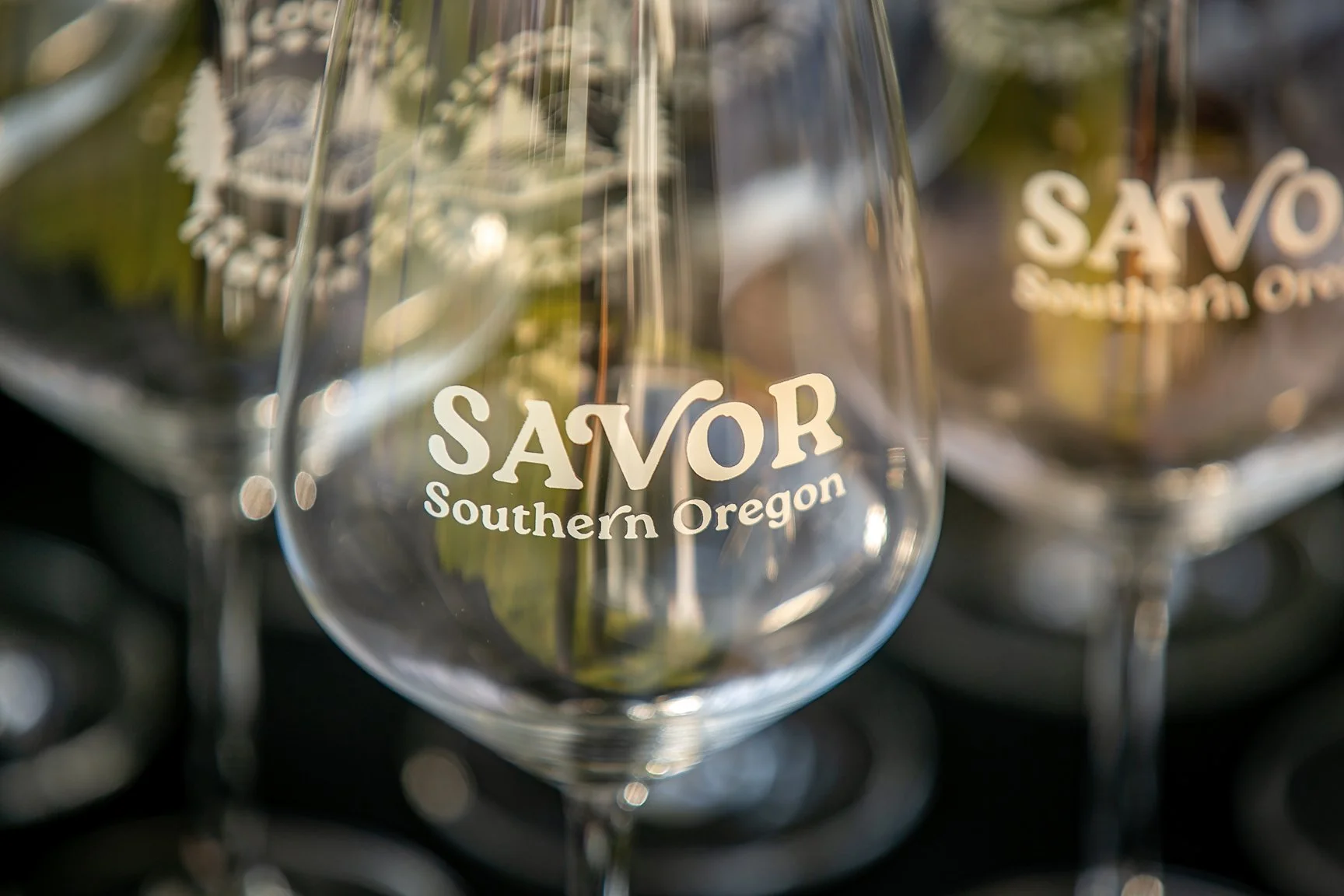 Close-up of a clear wine glass with the words 'SAVOR Southern Oregon' printed on it, with similar glasses blurred in the background.