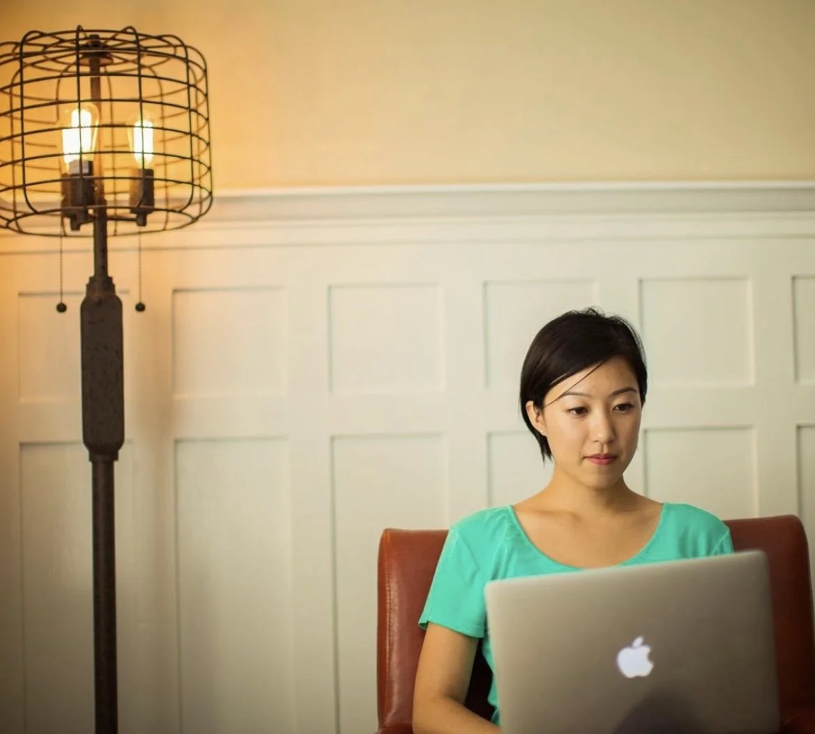 A woman with short black hair working on a silver MacBook in a room with beige walls, seated on a brown chair near a tall floor lamp with exposed bulbs.