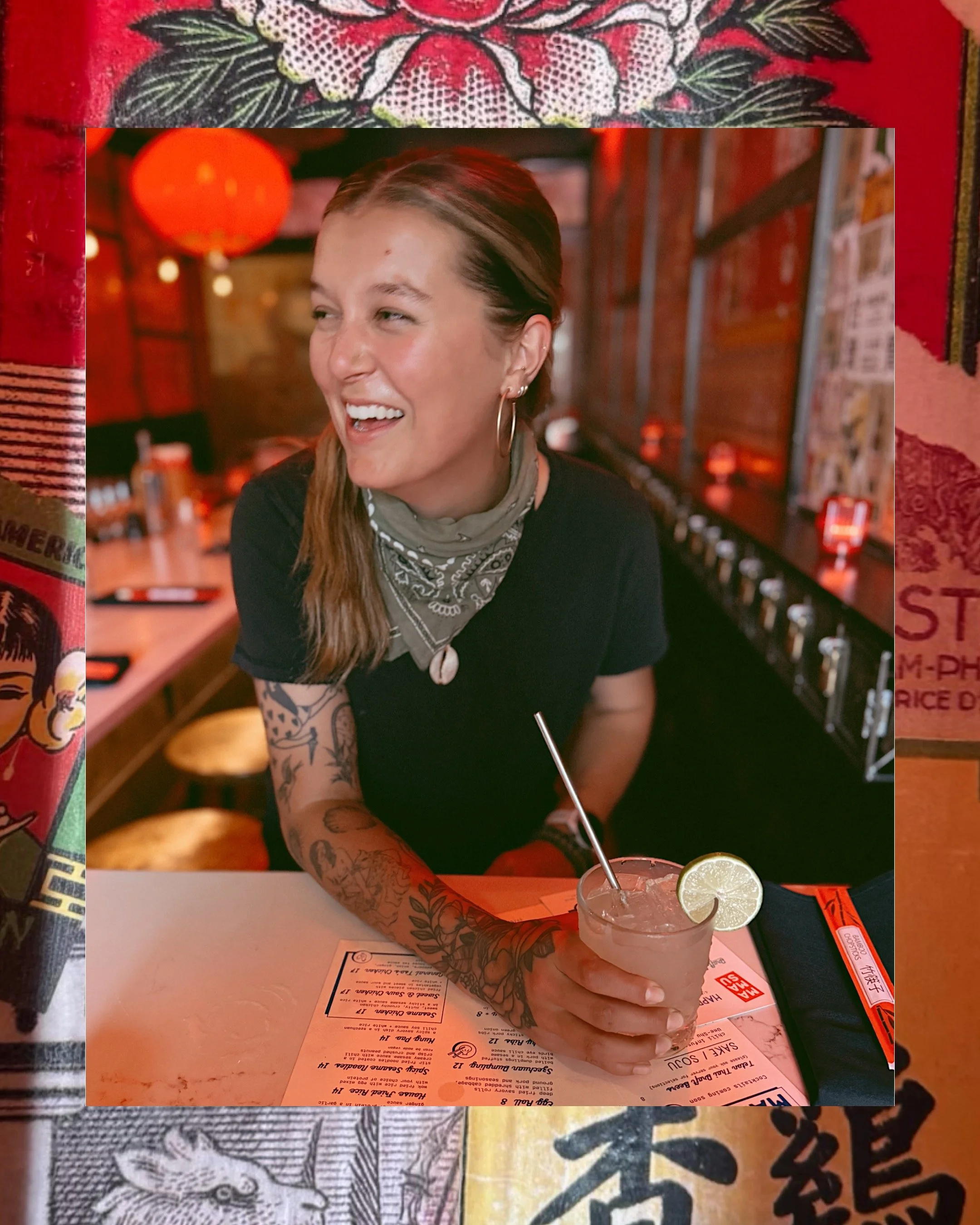 A smiling woman with tattoos on her arm, wearing hoop earrings and a bandana around her neck, sitting at a table in a dimly lit restaurant or bar, holding a cocktail with a lime wedge.