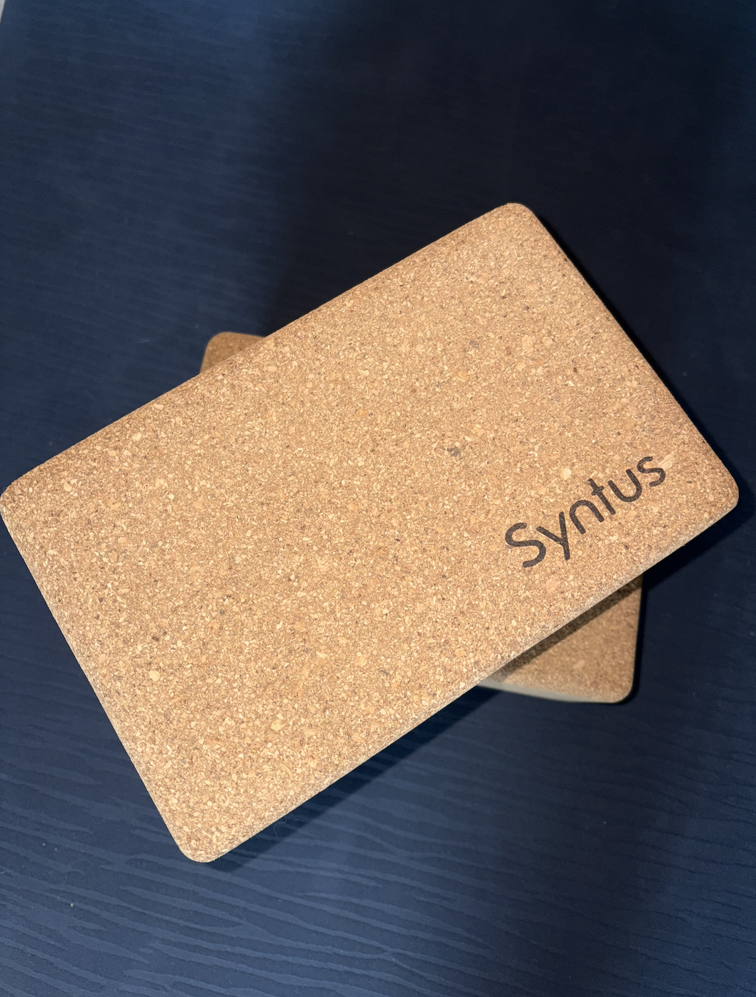 A rectangular cork yoga block with the word 'Syratus' printed on it, placed on a dark blue surface.