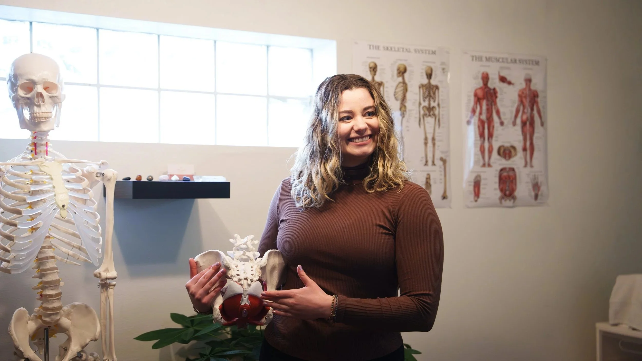 A woman standing in a classroom holds a model of a pelvis and smiling. Anatomical charts and a skeleton model are on the wall behind her.