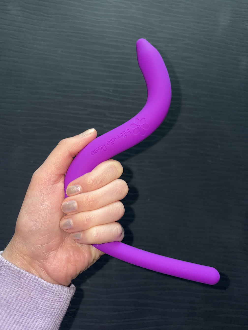 Hand holding a purple curved silicone pelvic therapy tool against a black wood grain background.