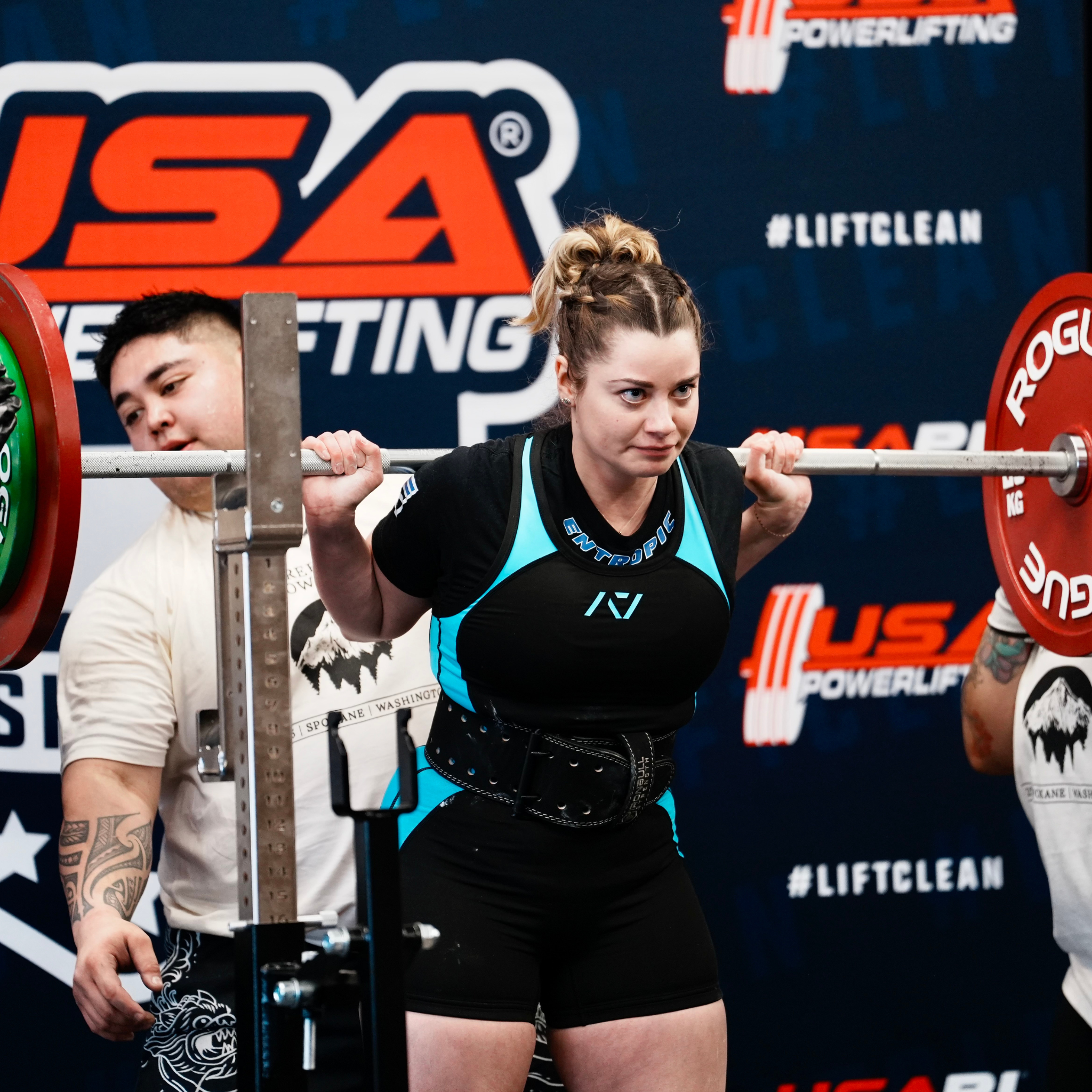 A woman lifting weights with a barbell during a powerlifting event, with a focused expression, in front of a U.S.A Powerlifting banner.