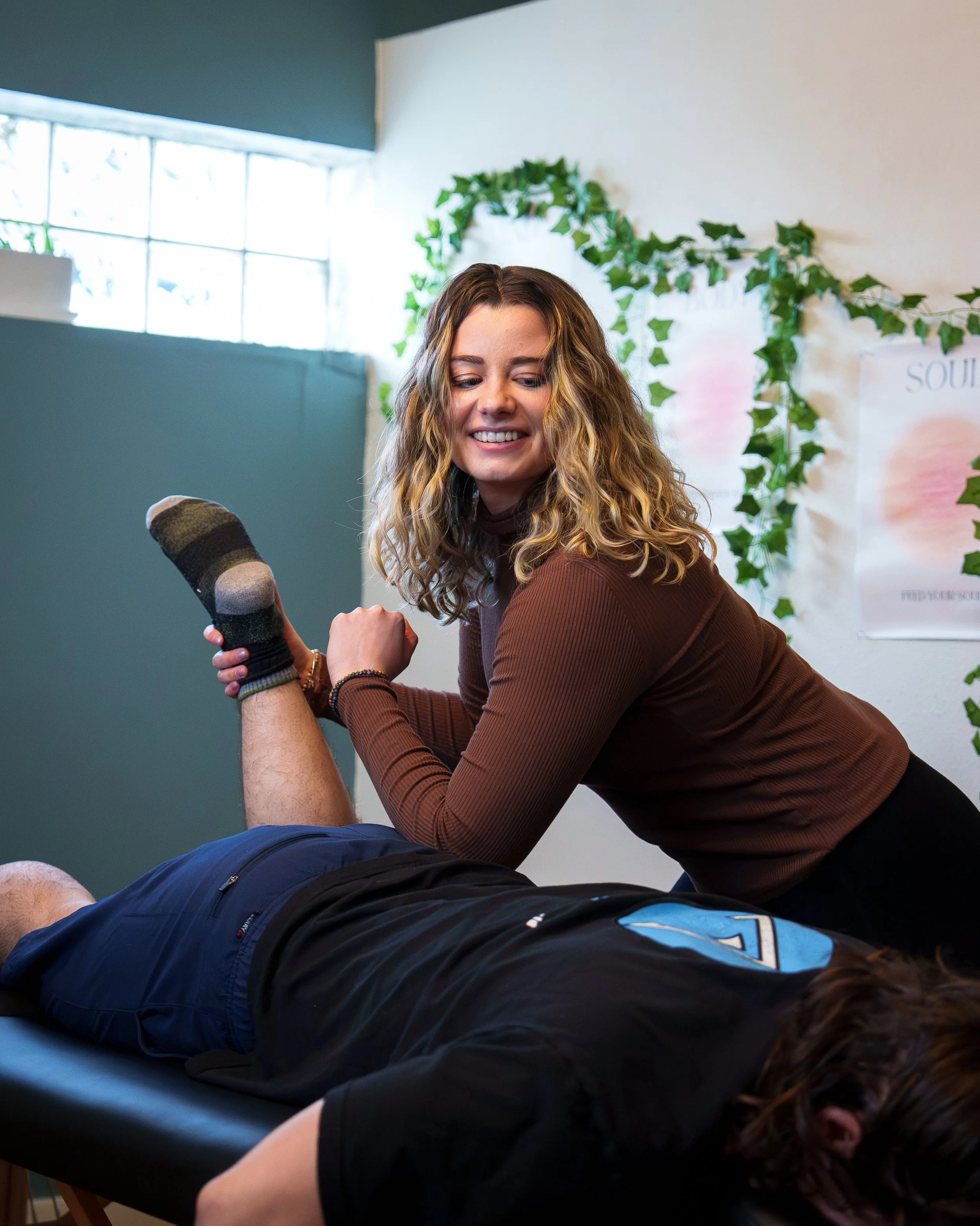 A woman in a brown long-sleeve shirt smiling while performing a physical therapy exercise on a person lying face down on a therapy table.