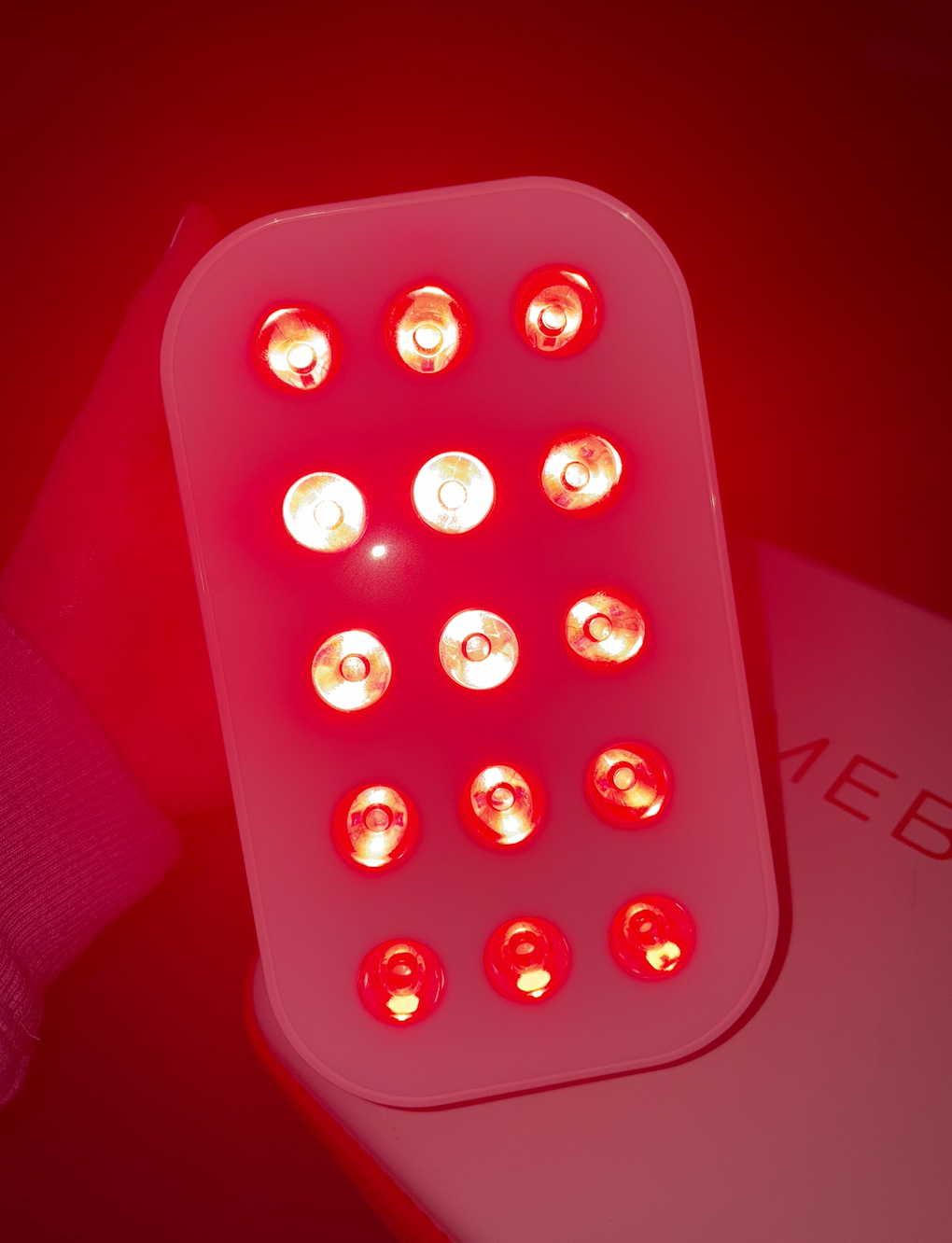 A handheld red LED light therapy device with multiple small glowing LEDs on a pink surface, emitting red light.