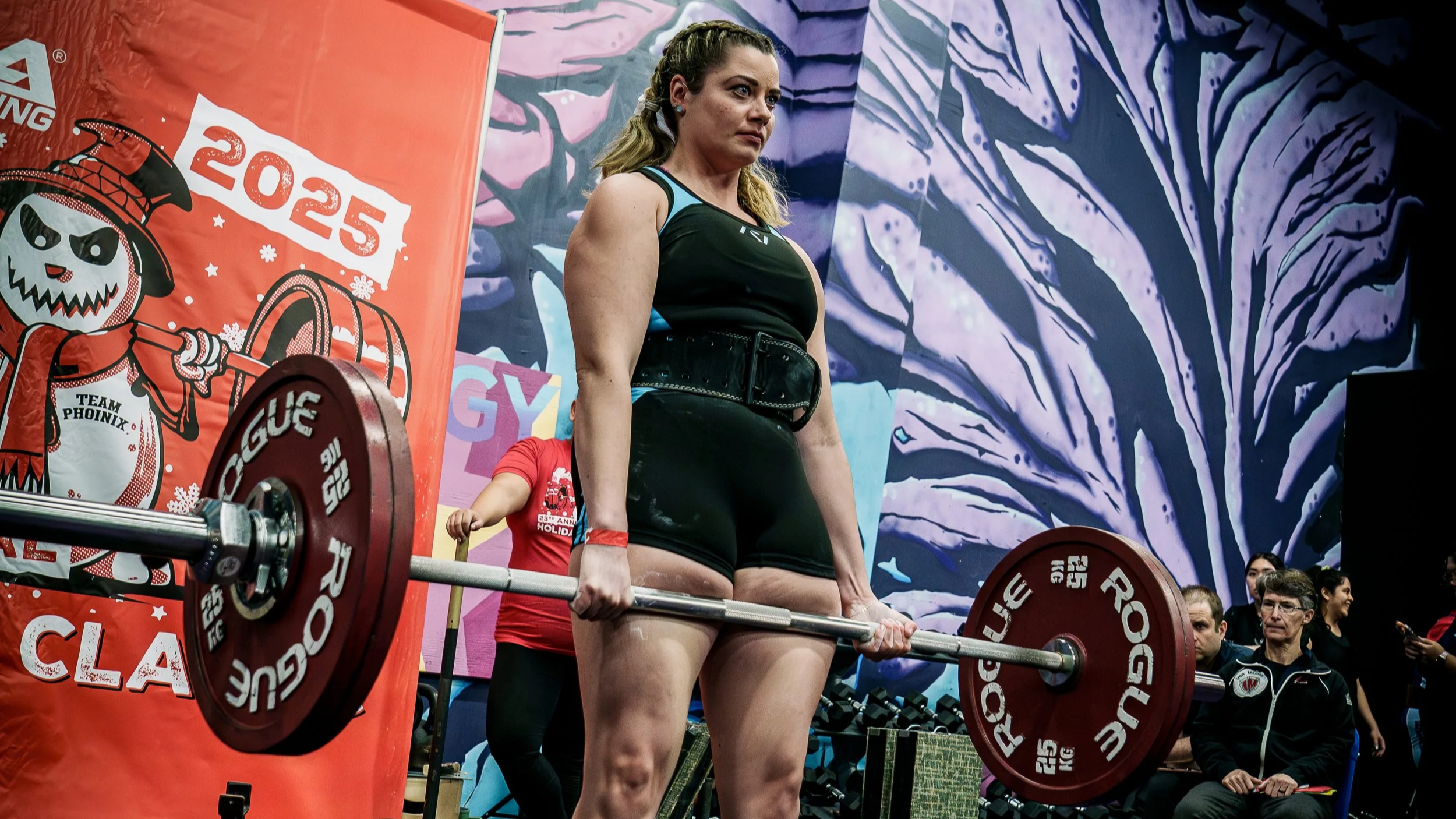 A woman with braided hair is preparing to lift a barbell during a weightlifting event, with an audience watching in the background.