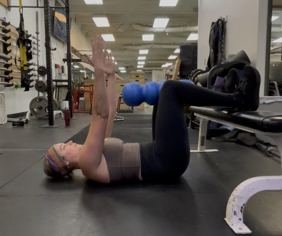 A woman lying on her back on a gym floor, holding a blue weighted ball above her chest, with her knees bent and feet flat on the floor, in a fitness or workout setting.