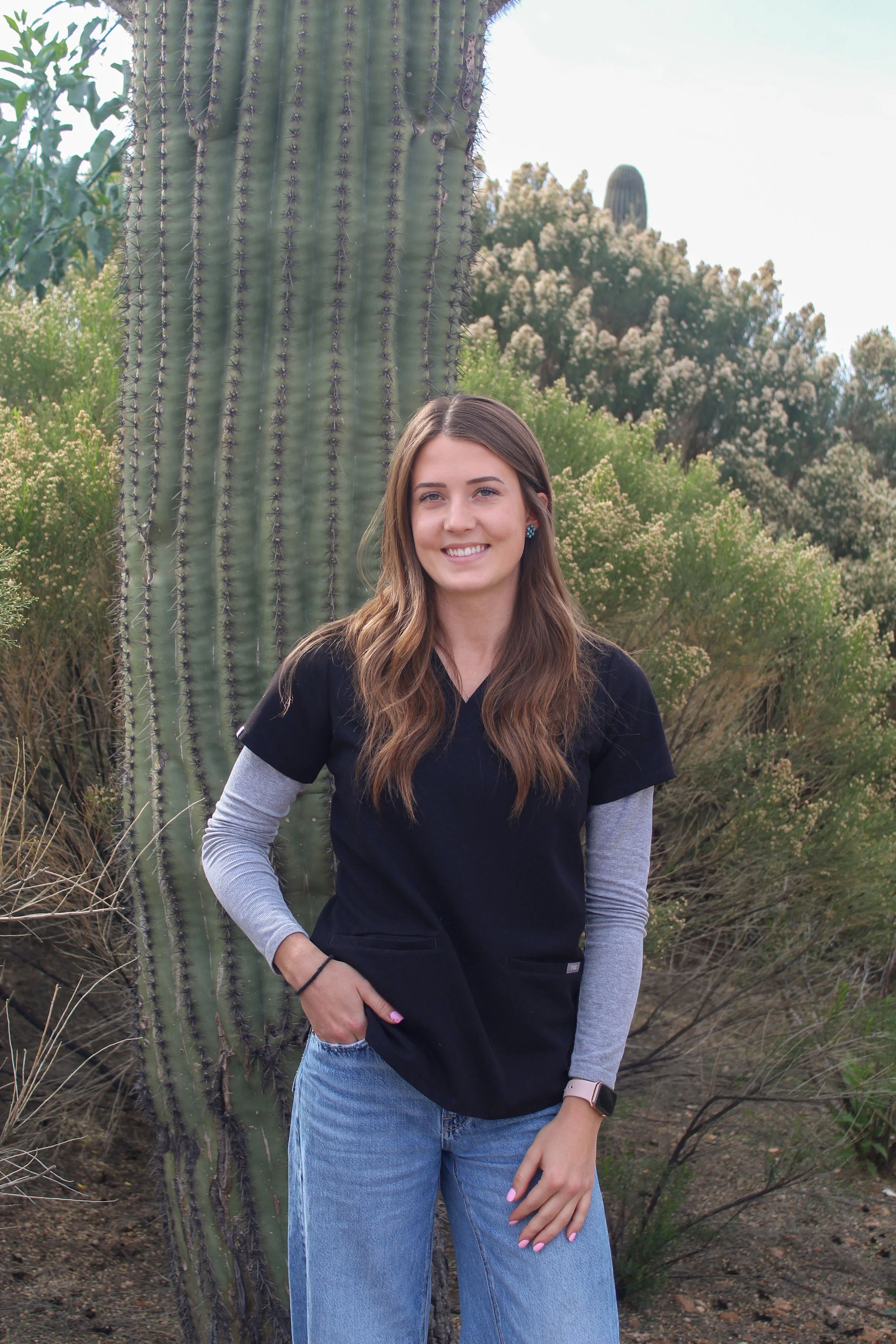 A young woman with long brown hair, wearing a black scrub top, gray long-sleeve shirt, and blue jeans, standing outdoors in a desert landscape with a large cactus and various desert plants in the background.