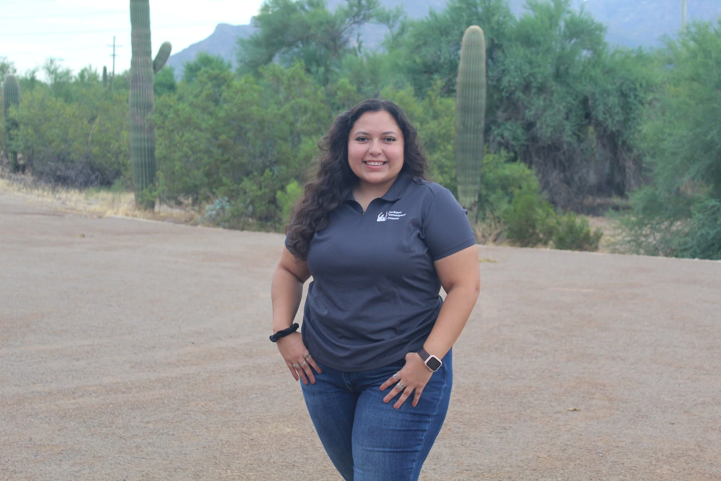 Smiling woman standing outdoors with desert plants and cactus in the background, wearing a gray polo shirt and jeans.