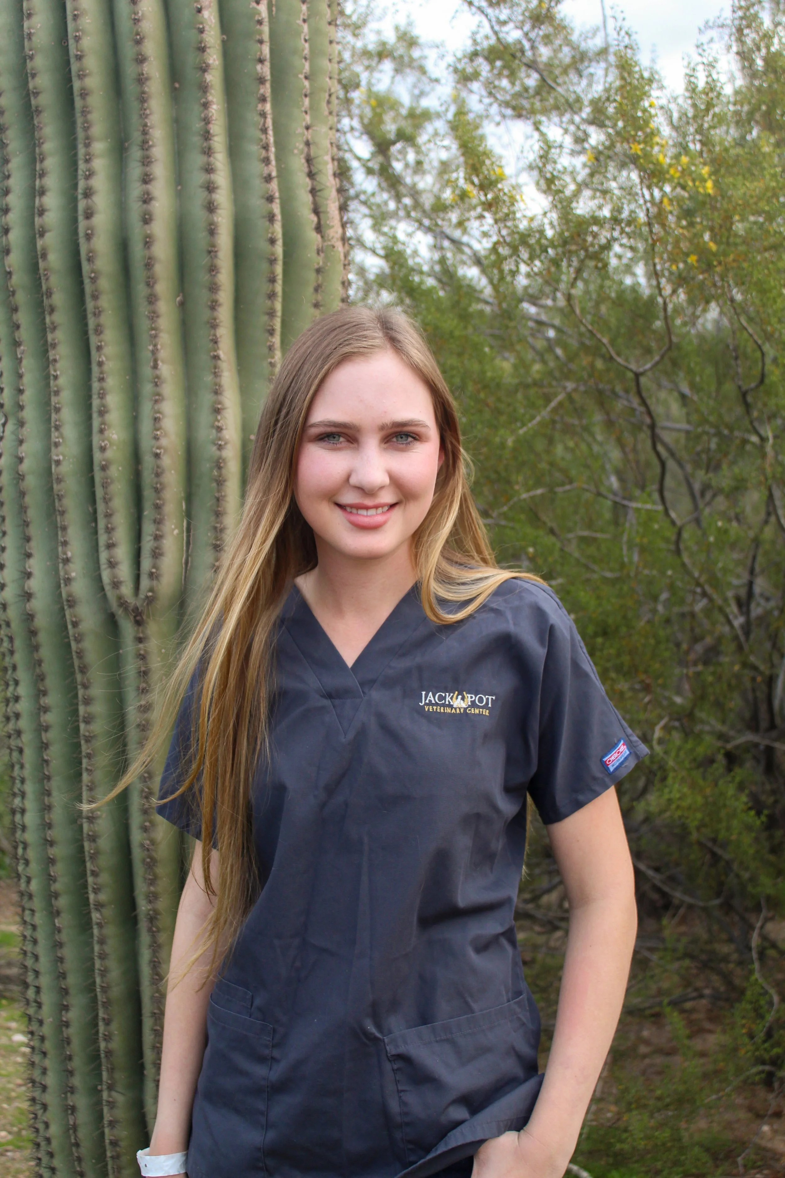 A young woman with long blonde hair smiling while standing outdoors next to a large cactus, wearing a navy blue veterinary scrubs top from Jackpot Veterinary Center.