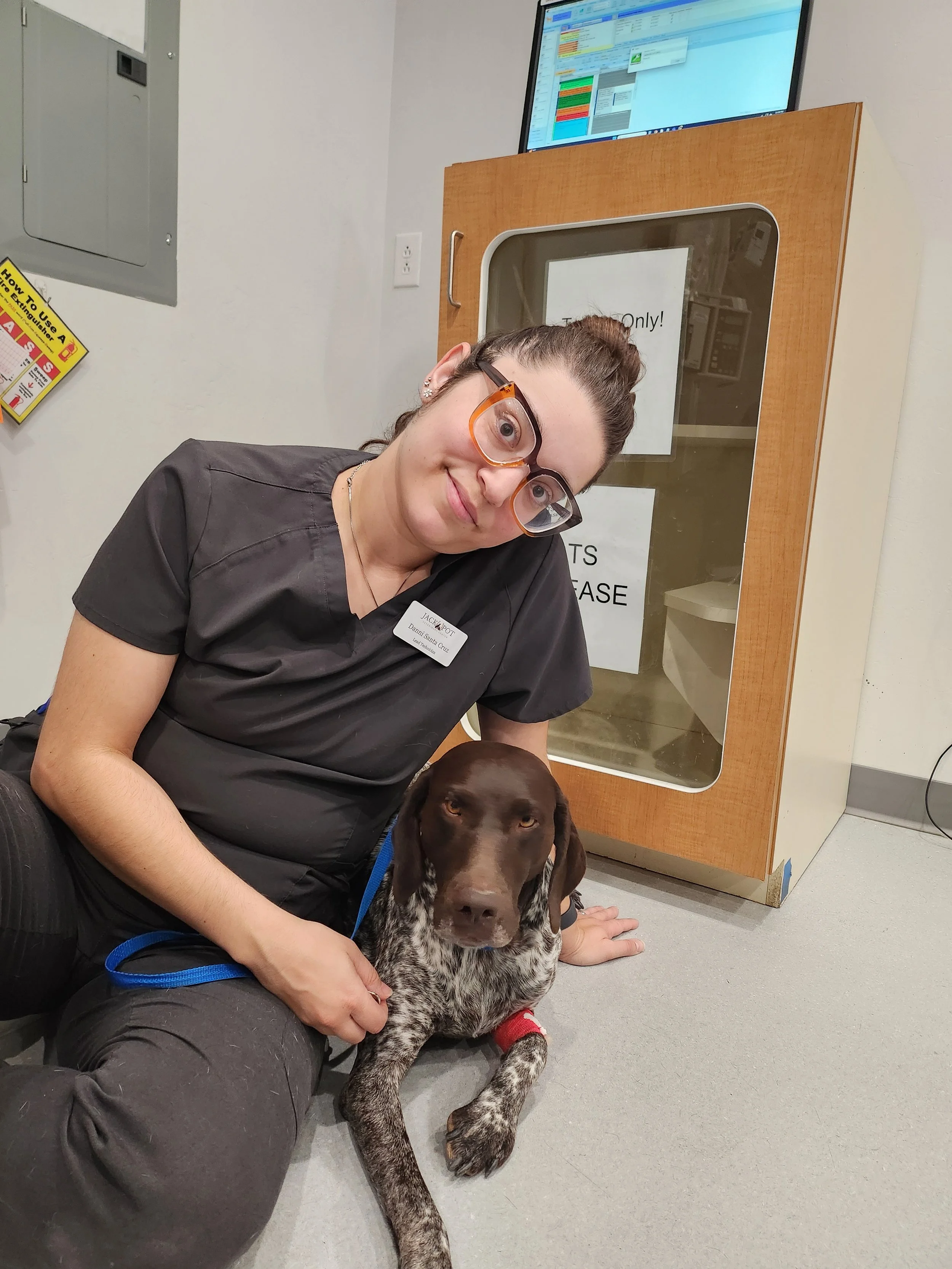 A woman in scrubs sitting on the floor with a brown and white speckled dog, against a background of a door with signs and a computer screen.